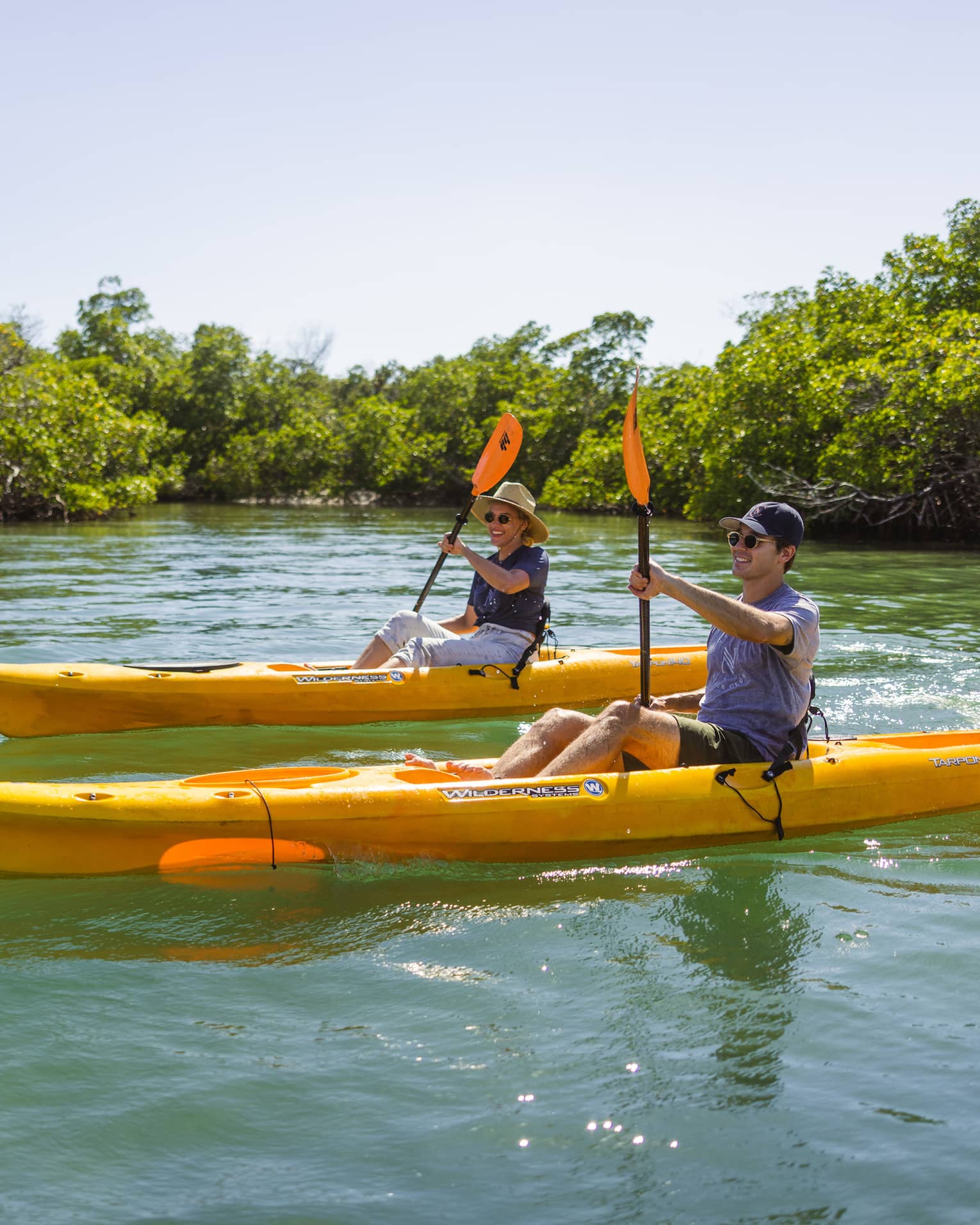 Two people row in yellow kayaks on the water, with bushy trees lining the waterway