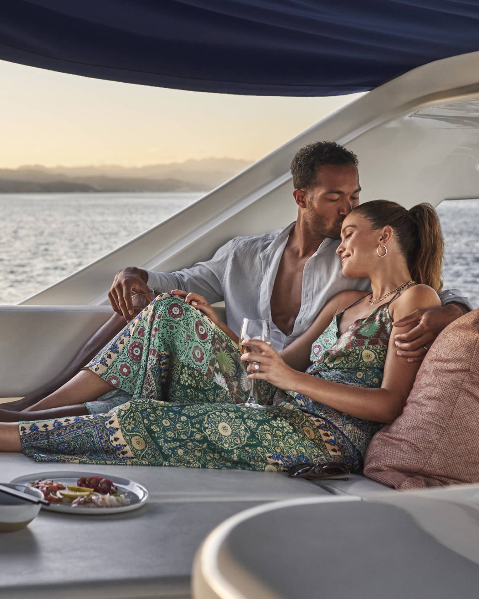 At sunset, a couple lounges in a luxury yacht set with a wine cooler, a plate of lemon slices and a dish of vibrant seafood.