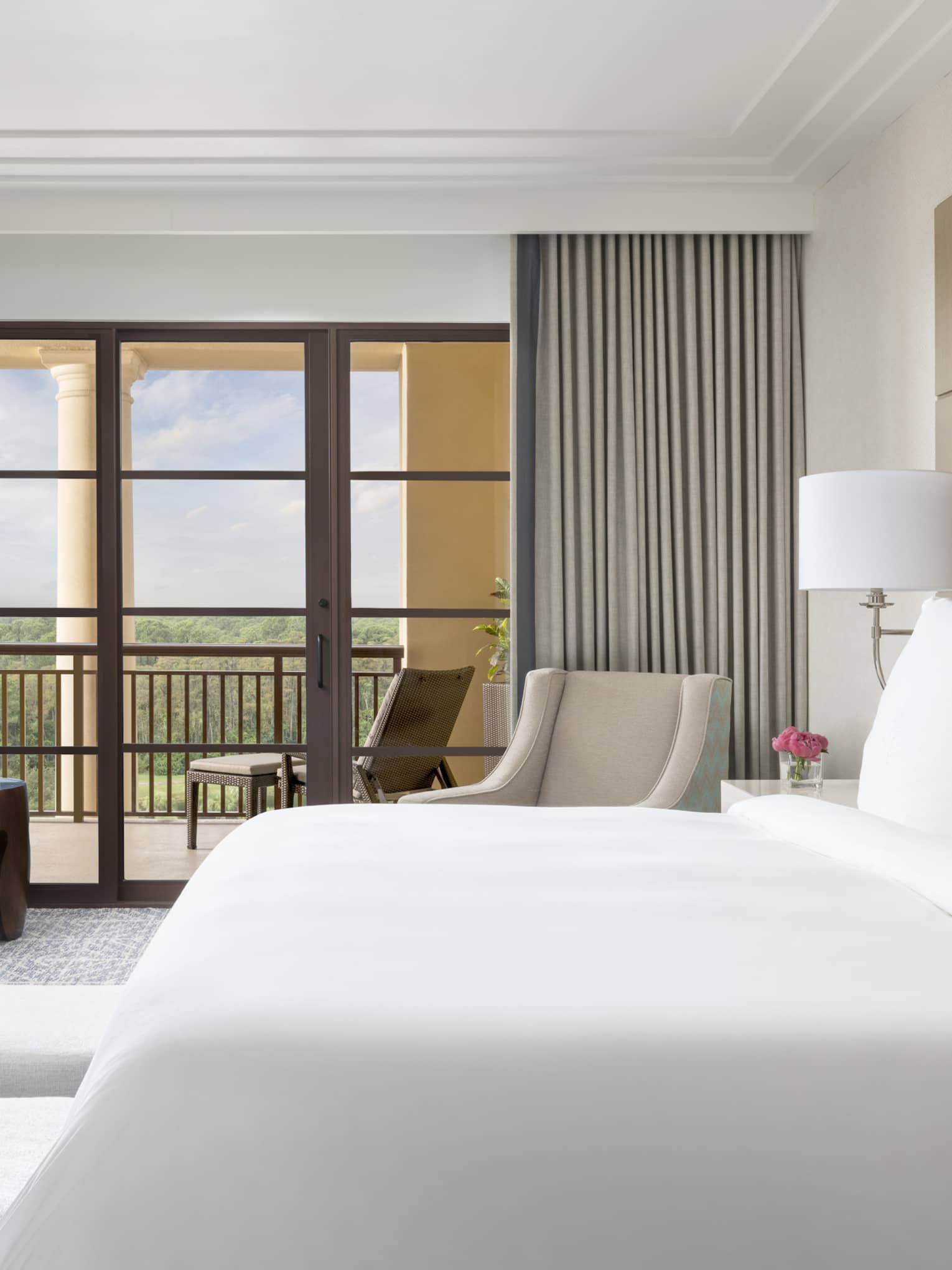 Hotel room with white king bed, two beige arm chairs, grey curtains, walk-out balcony
