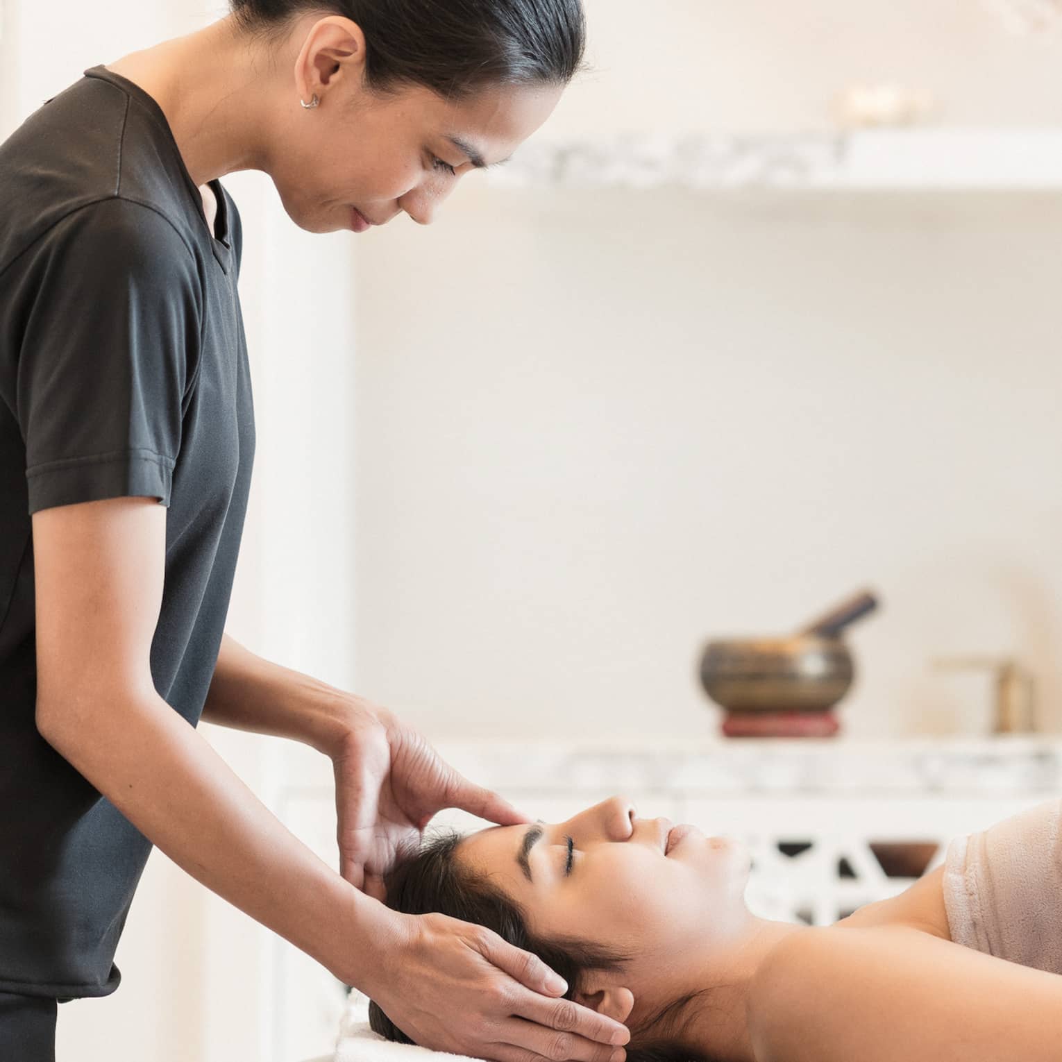 Massage therapist places hands on side of woman's face as she lays under a sheet on spa table