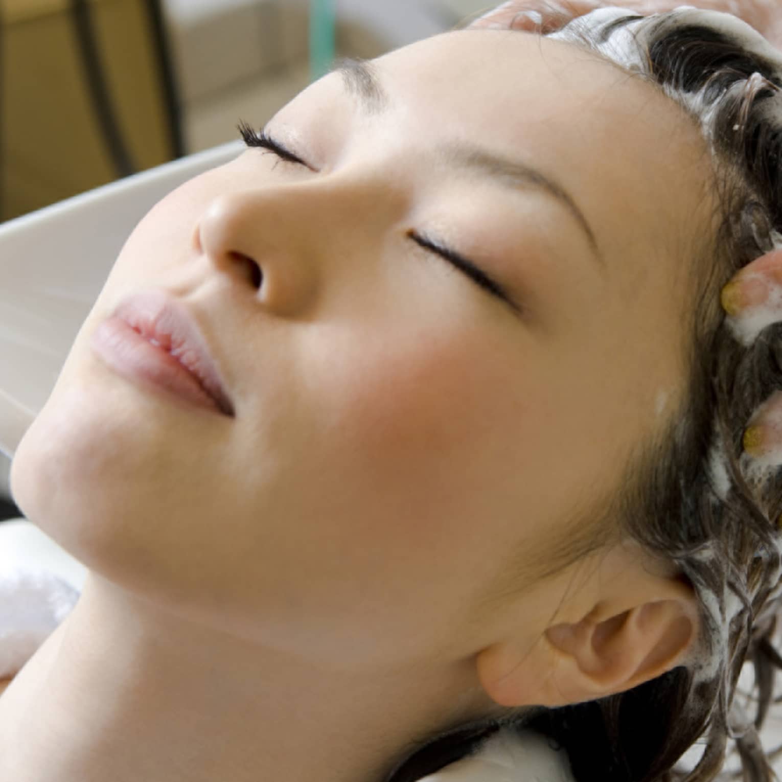 Woman closes eyes and leans back against sink as hands massage shampoo into her scalp