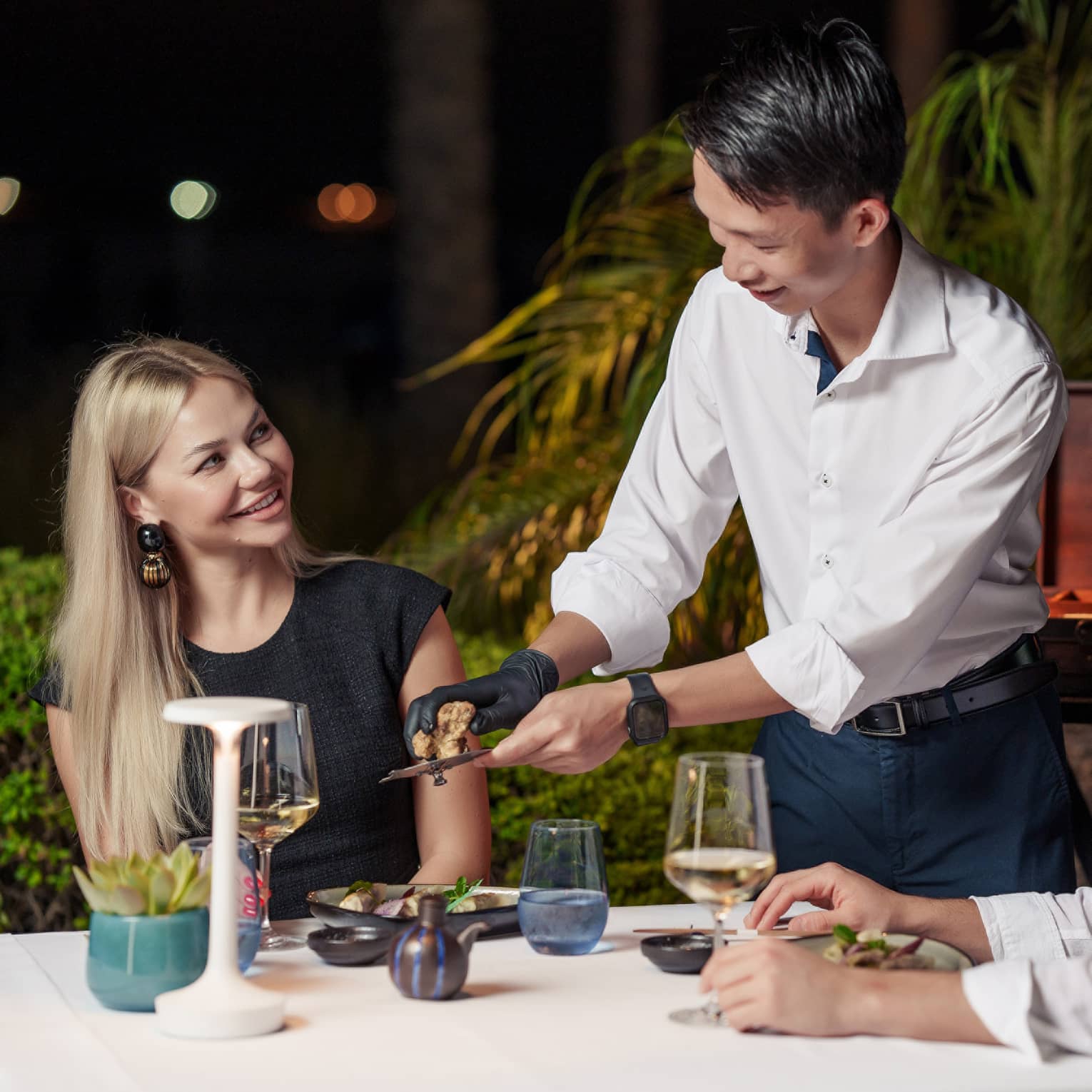 At a table for two amid foliage under the night’s sky, a server grates truffle over a diner’s dish as another diner looks on.