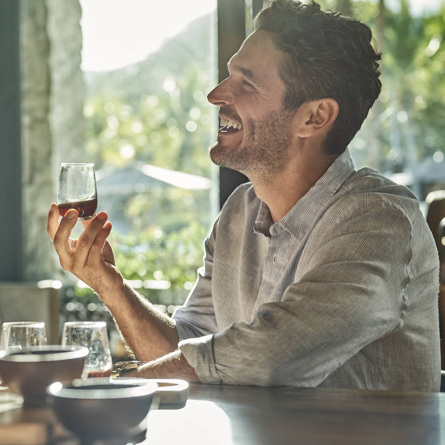 A man and a woman sit at a counter looking up and smiling and each holding a glass of rum with various other glasses and ingredients sit in front of them on the counter