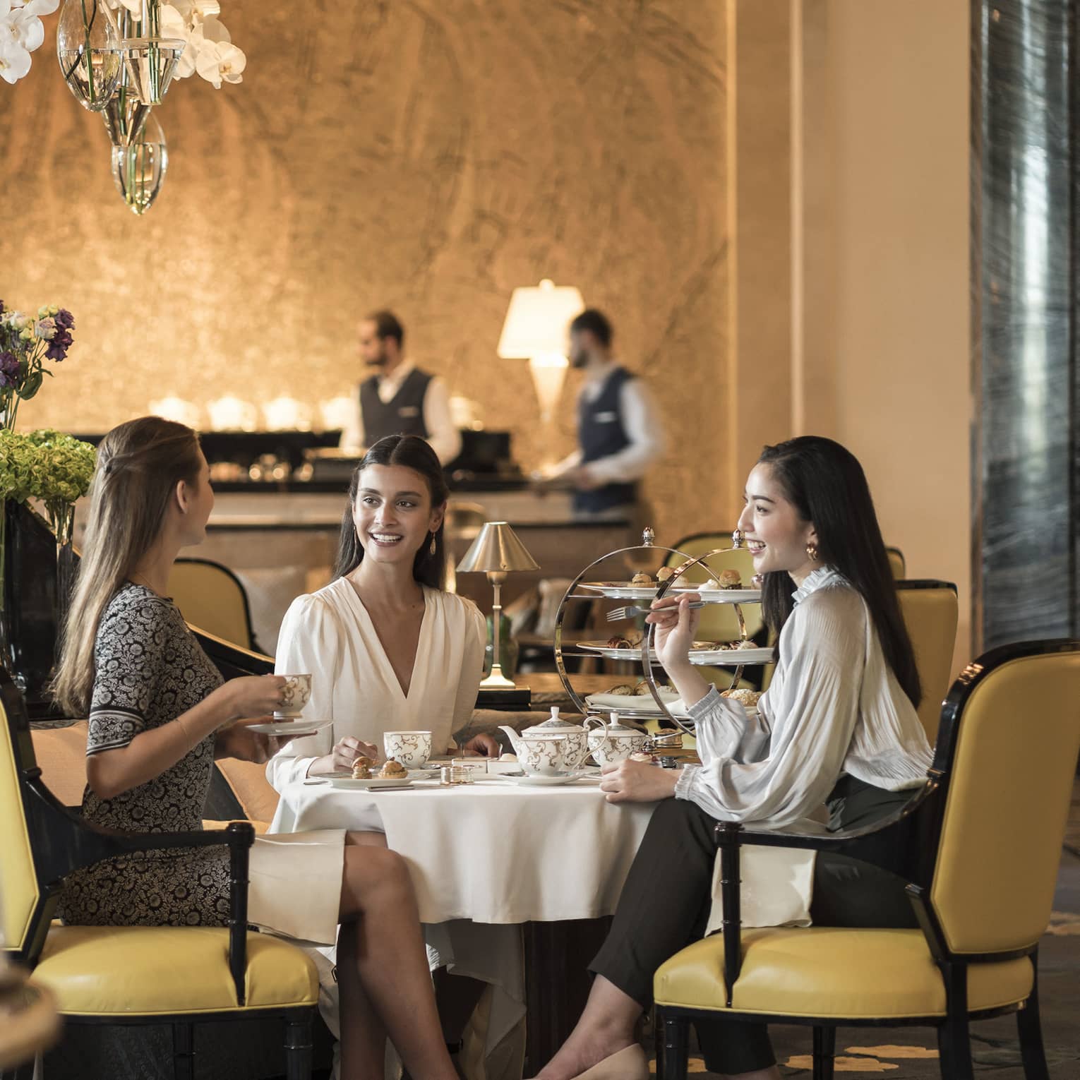 In an elegant restaurant, three smartly dressed diners enjoy an afternoon tea beside a feature of tiered flower-filled vases.