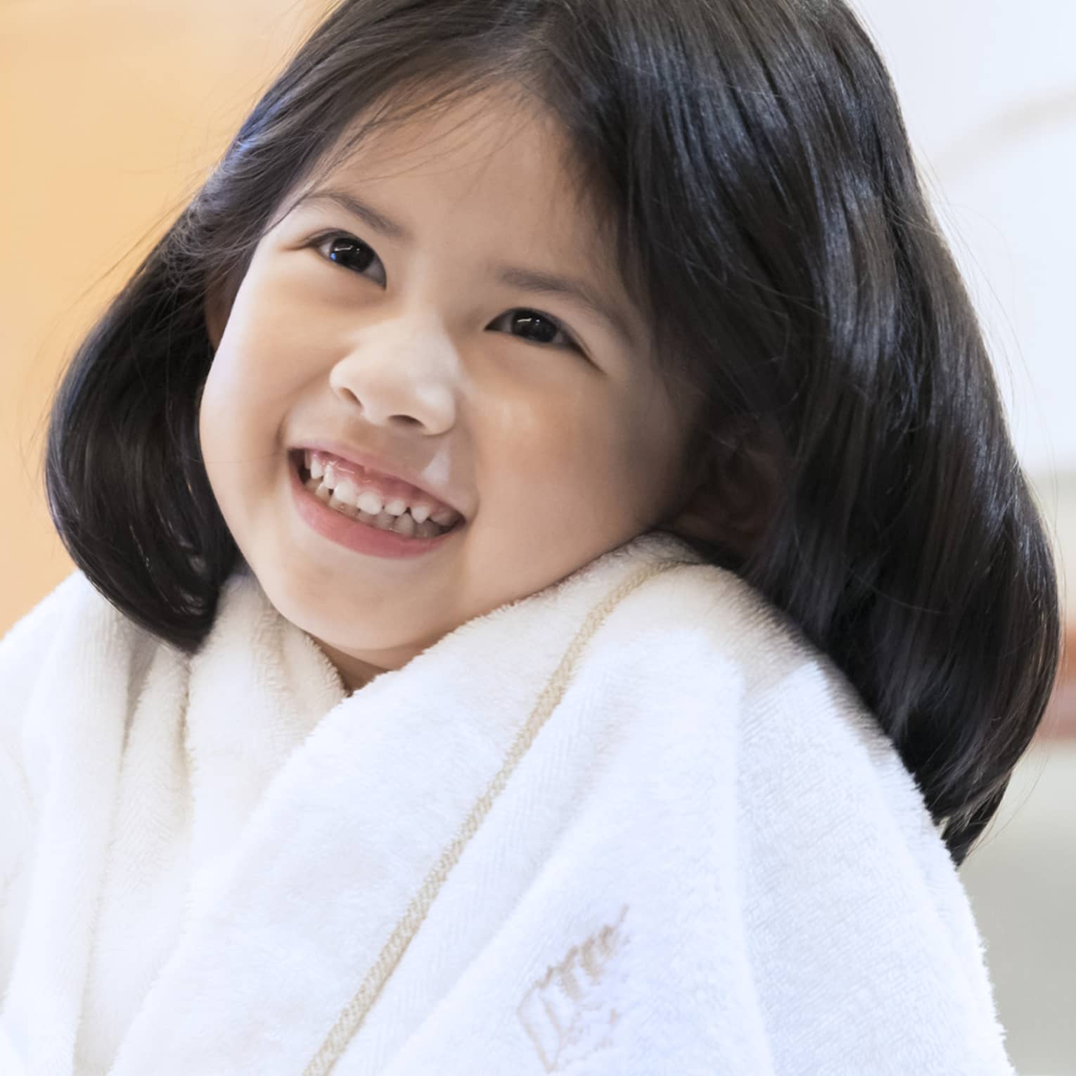 Smiling little girl wearing white bath robe and pink slippers sits on bed