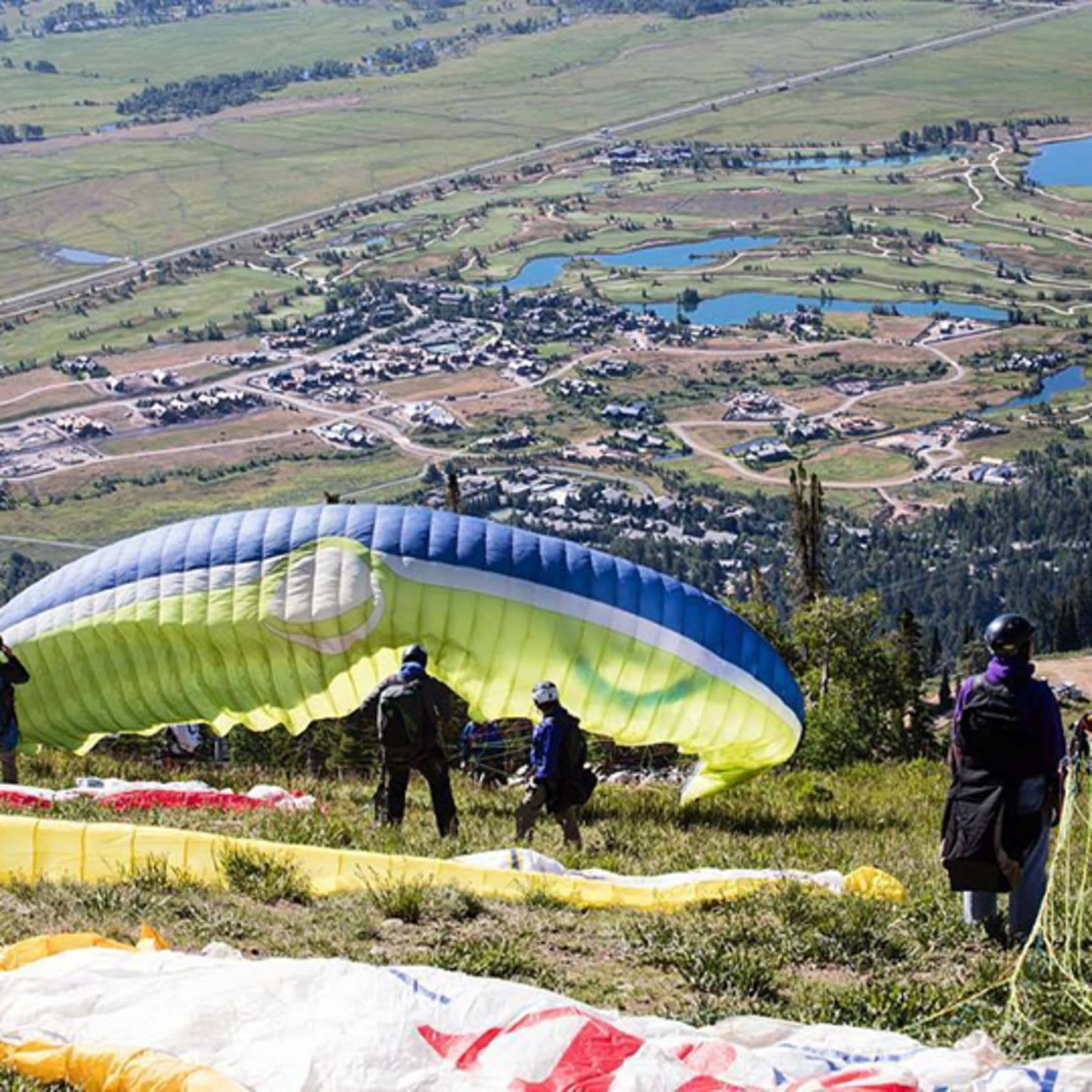 Paragliders preparing for takeoff on a grassy slope overlooking a valley with ponds, houses and open fields below