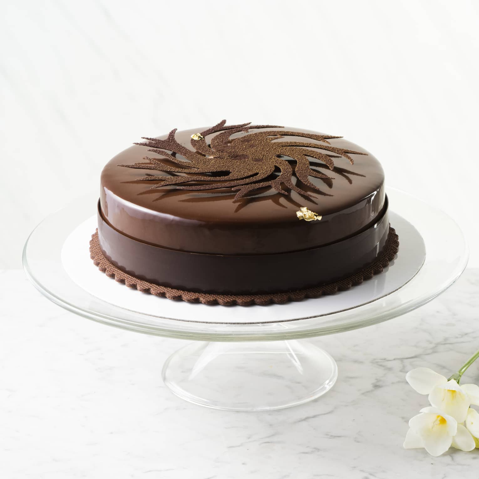 Round dark chocolate cake on glass cake plate and daffodils on table