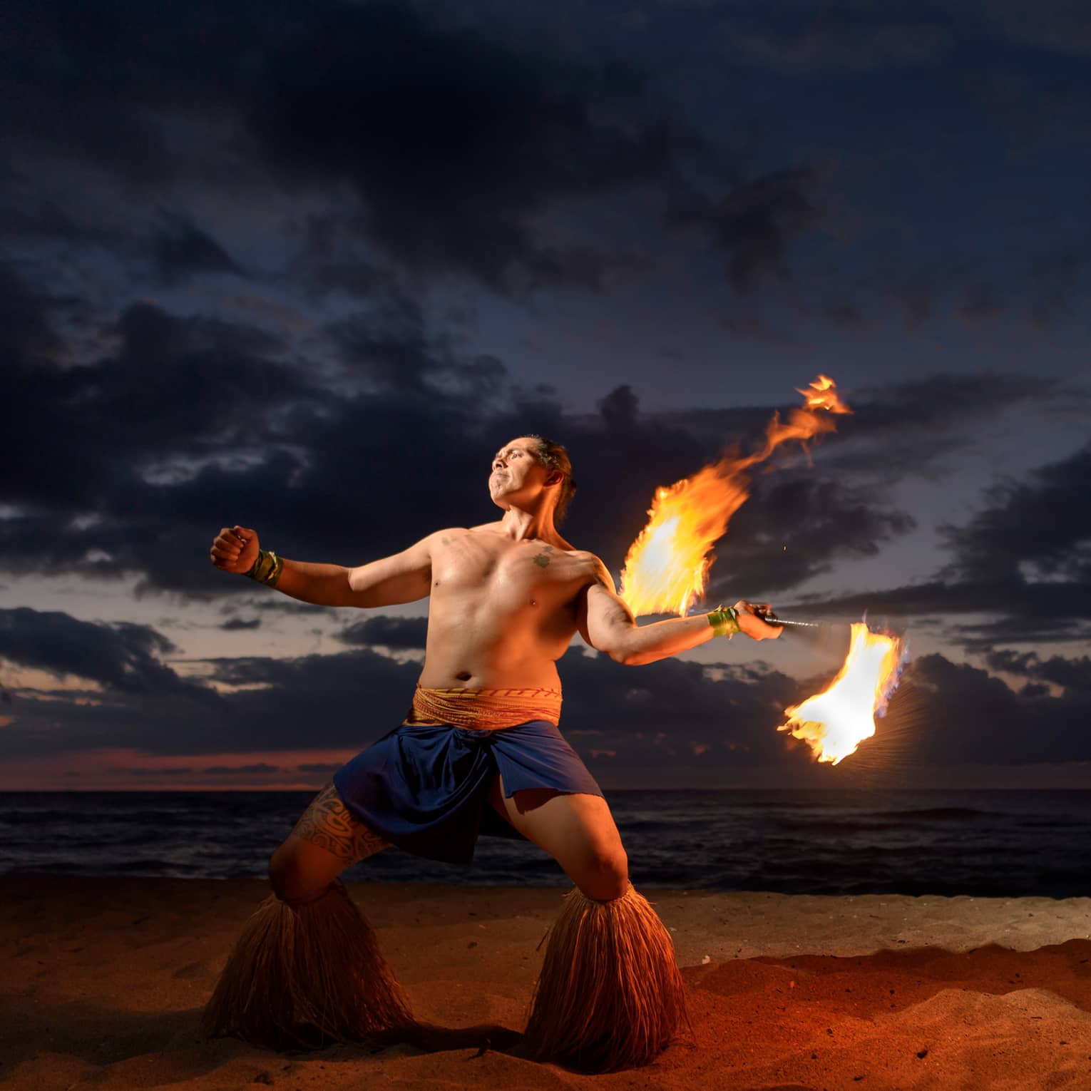 Man wearing traditional Polynesian outfit dances with torch on beach at night