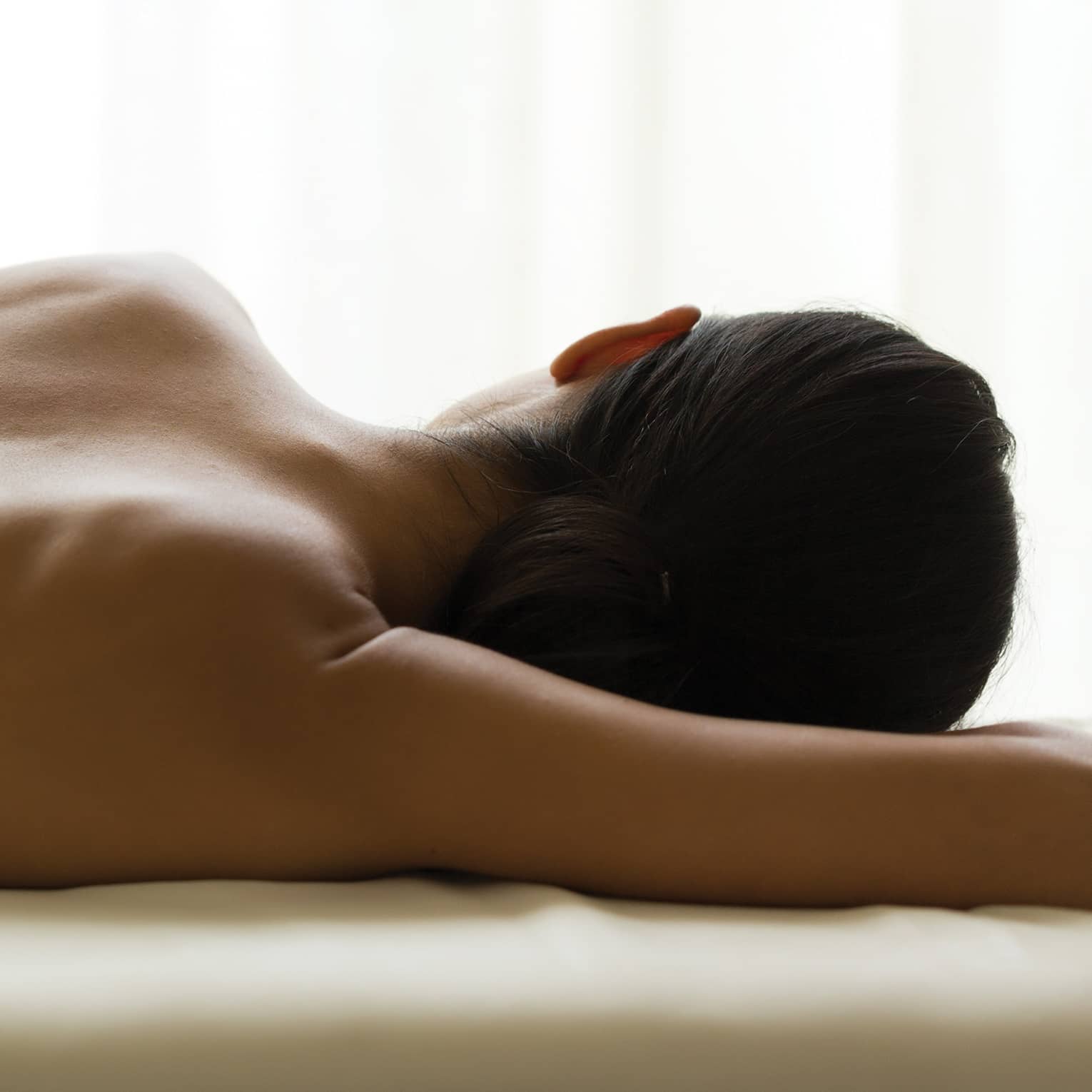 Woman's bare back as she lays on massage table under window, white curtains