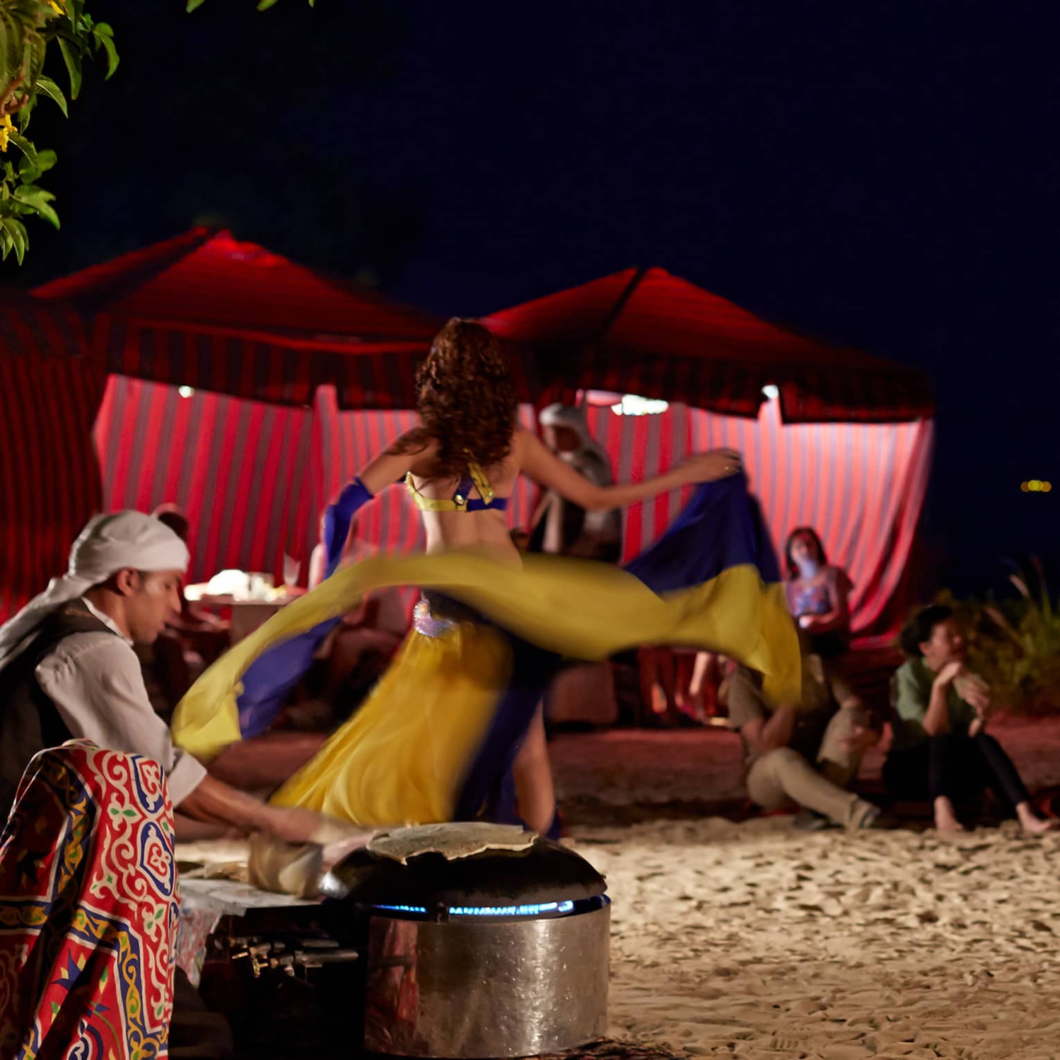 At night, a dancer performs outdoors for guests, swirling their skirt to music played by three smiling, seated musicians.