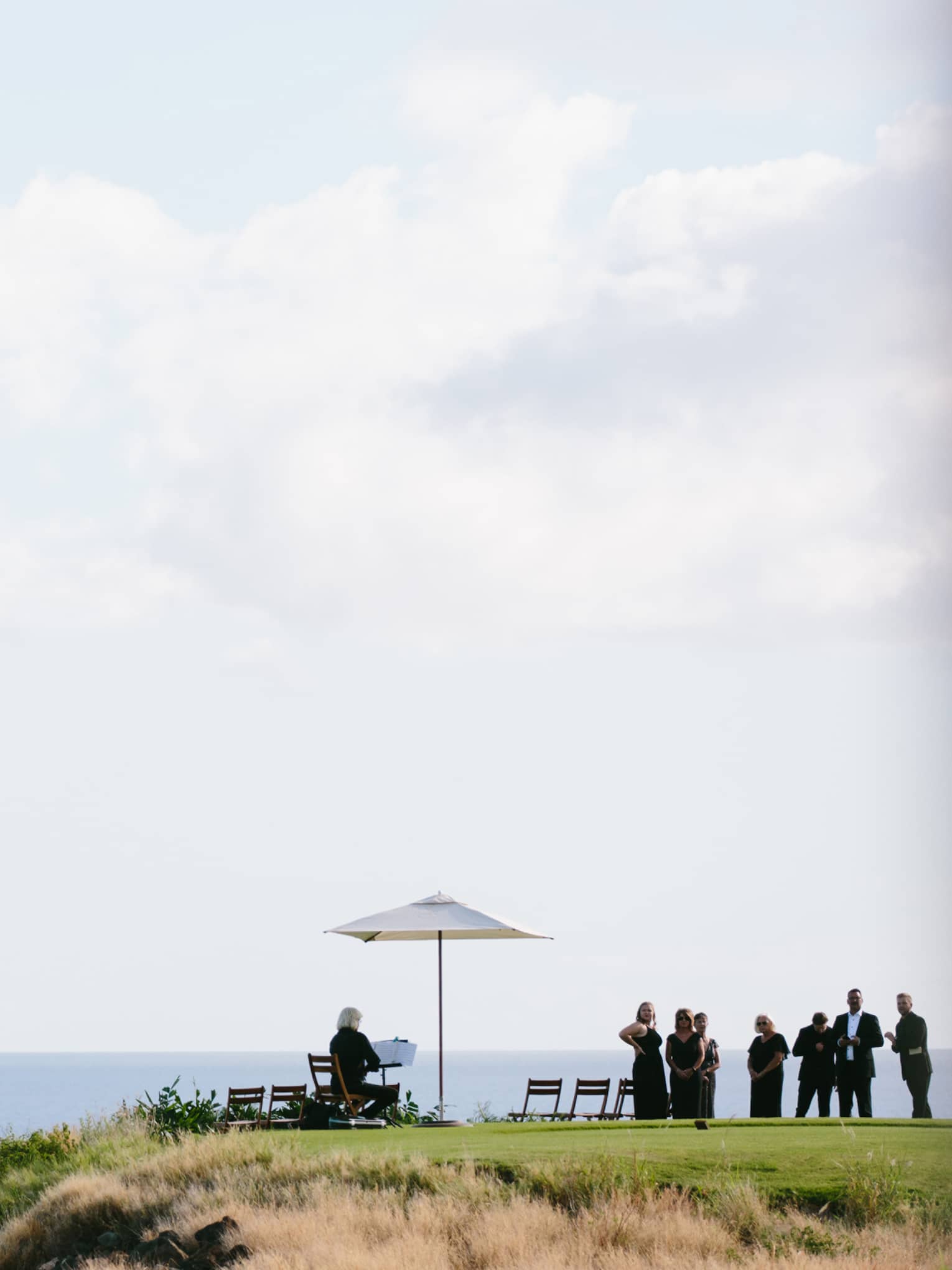 A group of people dressed in black congregate on the edge of a cliff overlooking a calm sea.