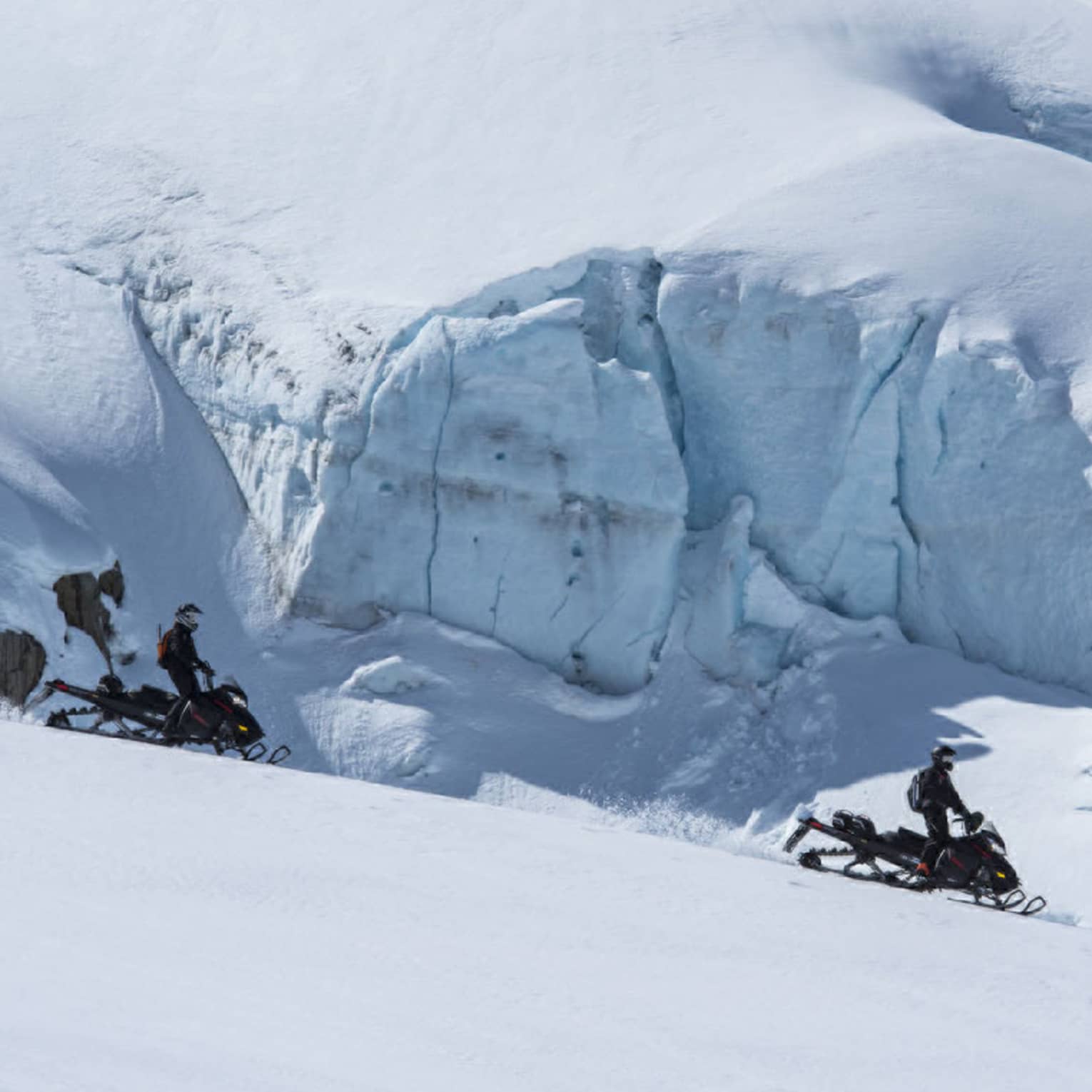 Side view of two distant black-clad snowmobilers coasting down a white slope alongside an enormous wall of ice and snow.