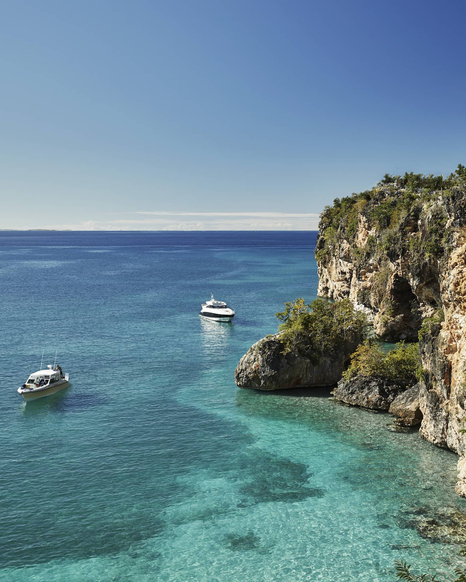 Long view of two cabin boats floating in the crystal-clear ocean beside towering rocky cliffs, a pale blue sky above.