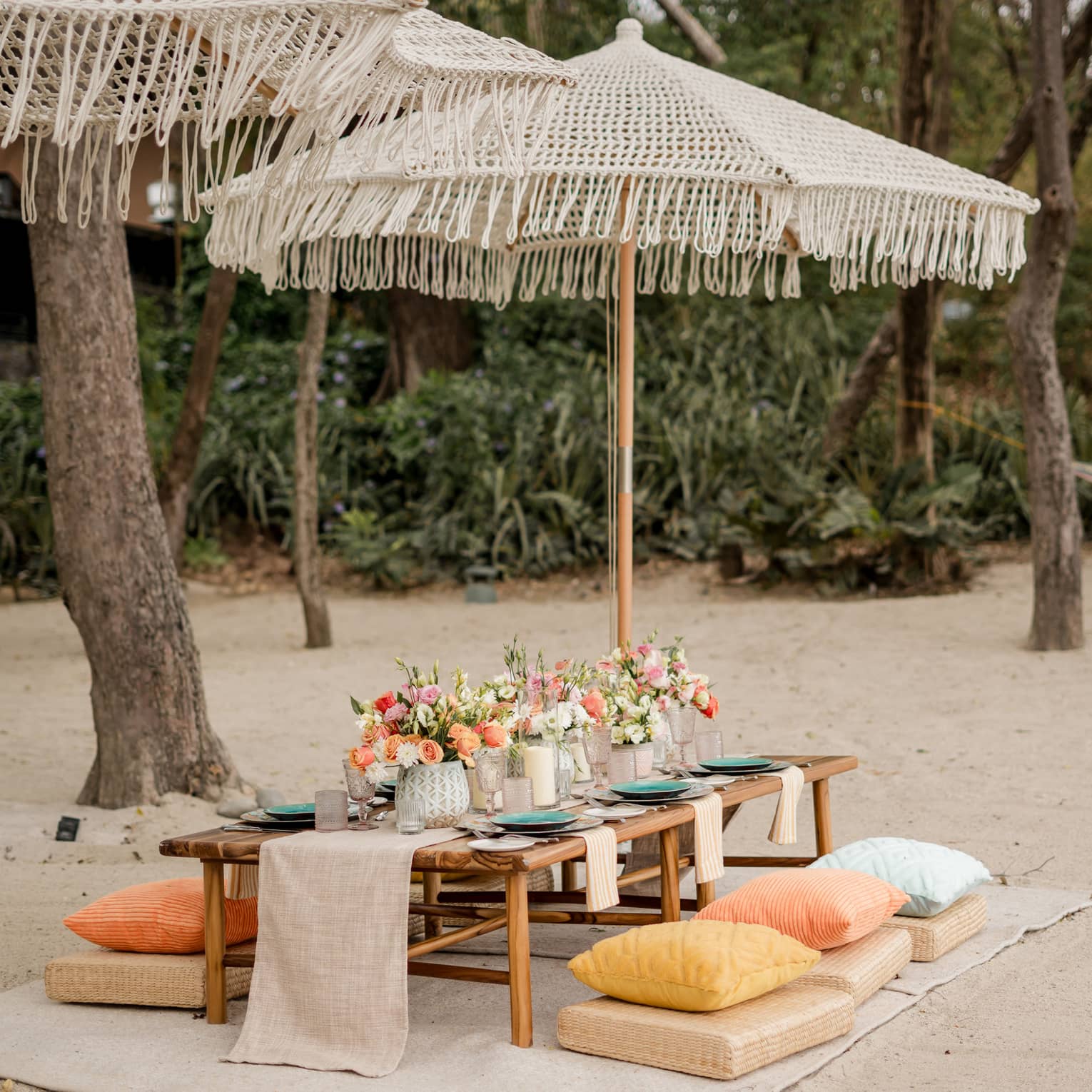 Wooden dining table set up with floral centrepieces and candles beneath an umbrella on a beach