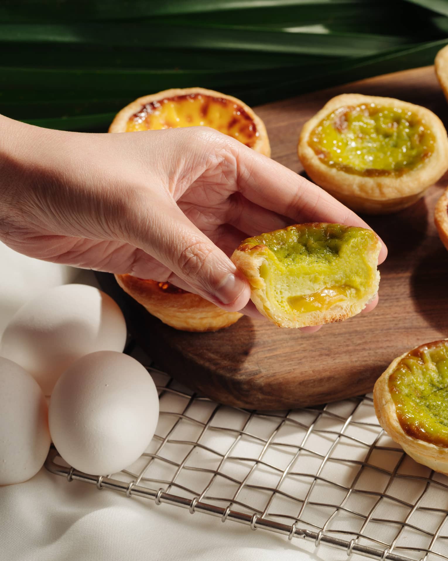 A hand holding a half-eaten Portuguese egg tart with a green filling, set against a backdrop of whole and partially-eaten egg tarts on a wooden board.