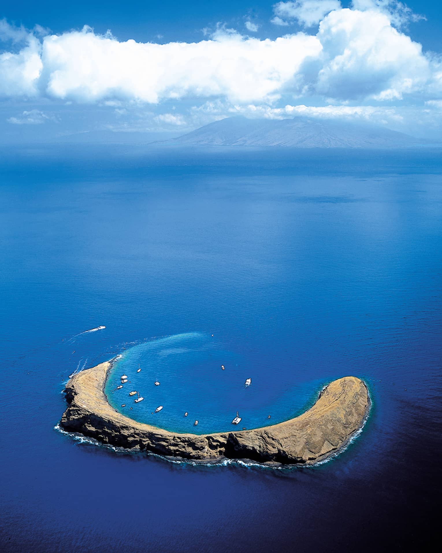 Aerial view of Molokini Crater, a crescent-shaped volcanic islet surrounded by clear blue ocean waters, with boats anchored nearby