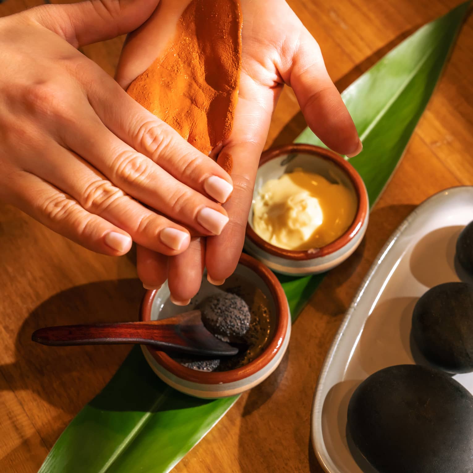 Pair of hands tests spa ingredients from two small stoneware bowls set on a fresh green leaf with three black stones placed on a dish nearby