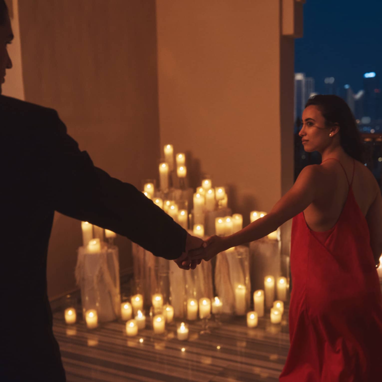 Person wearing a red dress leads another person who is silhouetted in the shadow by the hand toward a terrace filled with lit candles at night