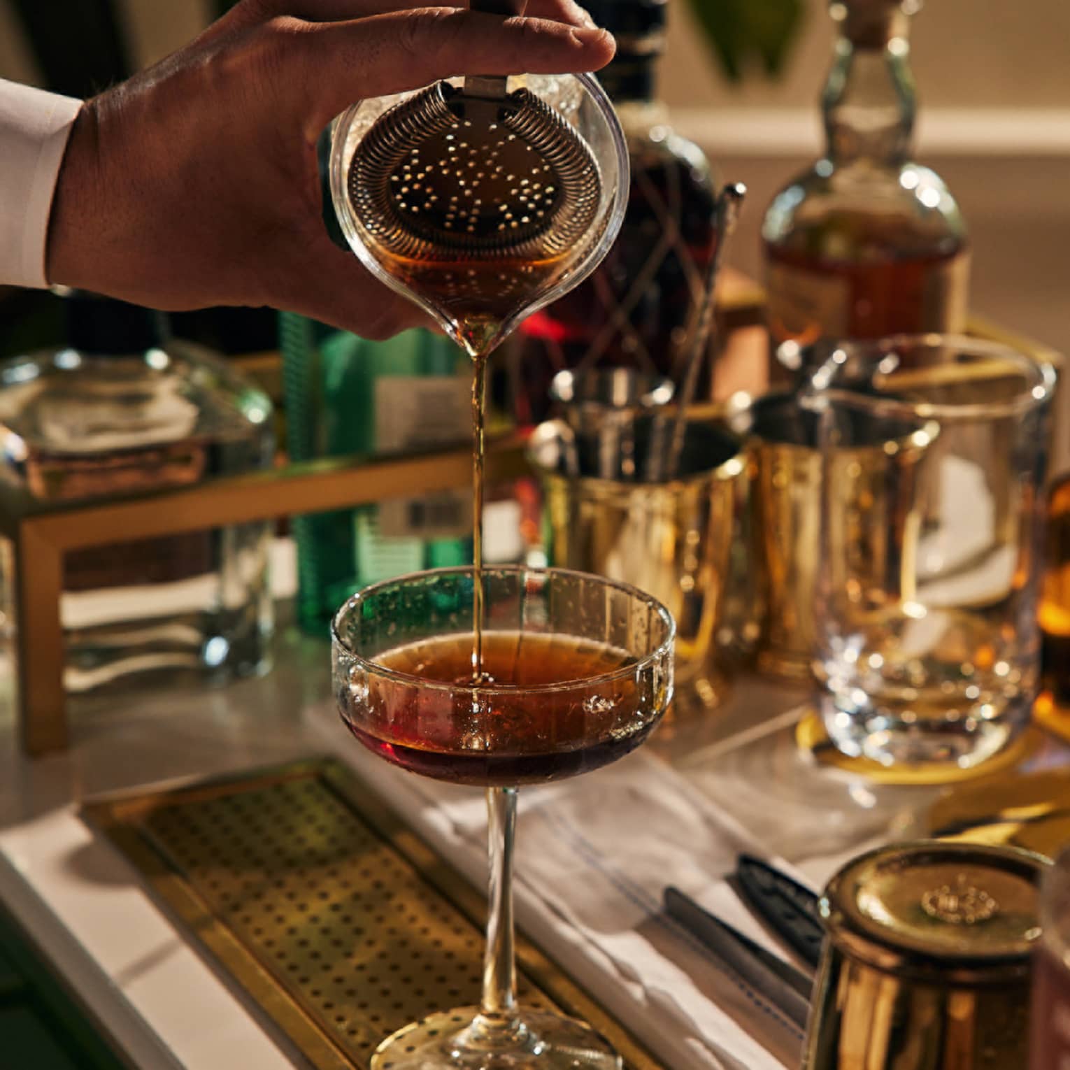 A bartender pouring a drink into a cocktail glass with bottles of liquor around.
