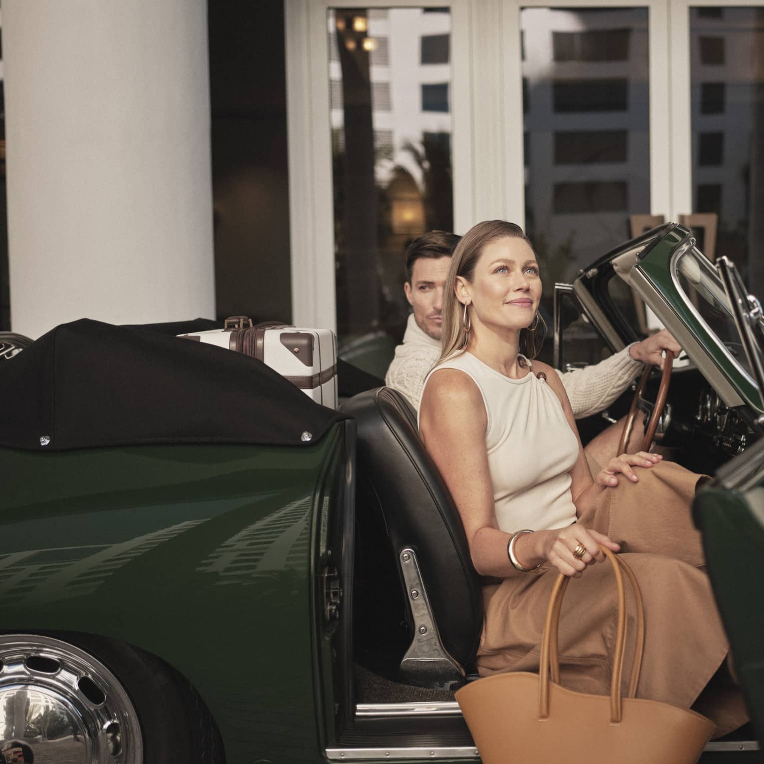 A man and woman getting out of a dark green car holding bags, two hotel team members are assisting them.