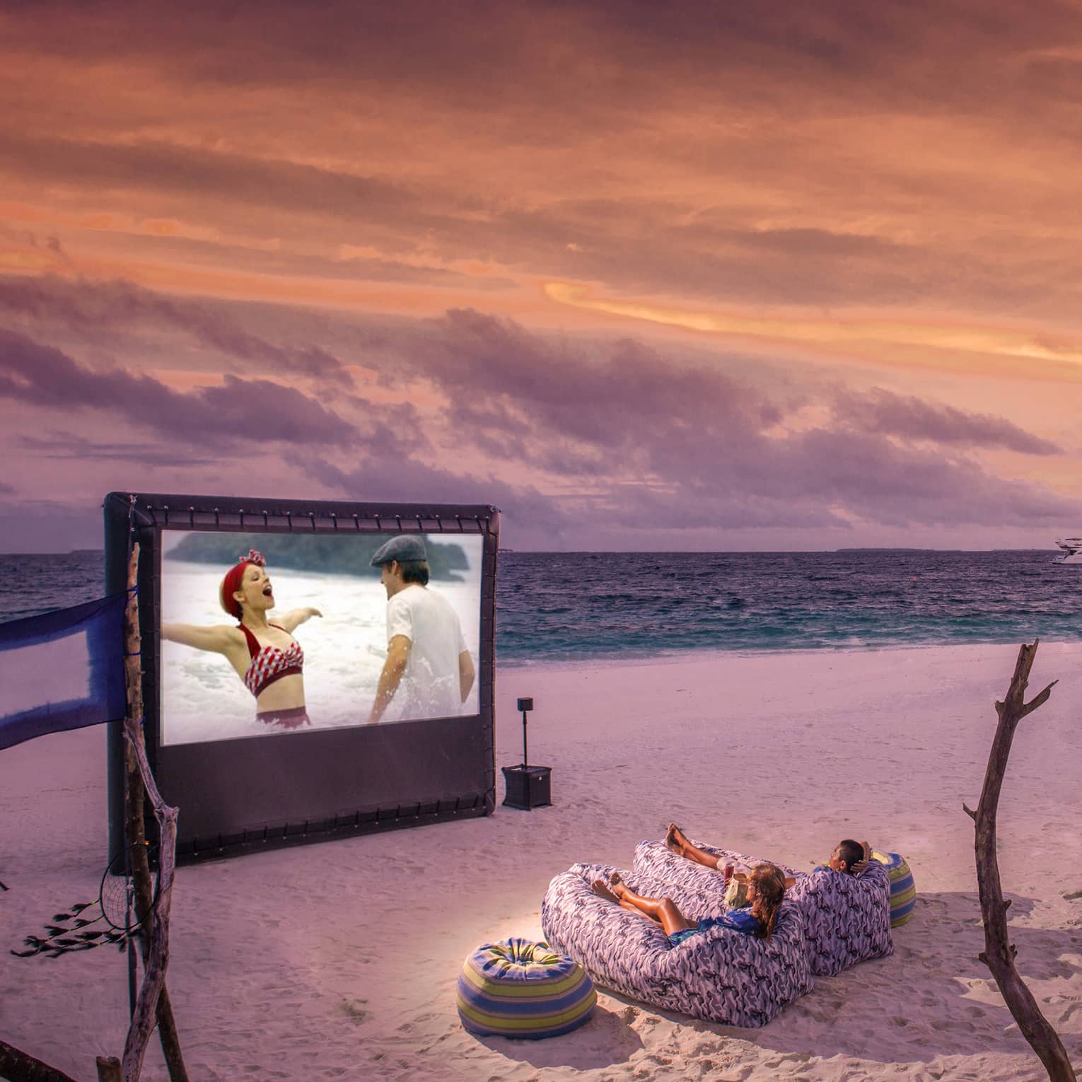 ,Two people in beanbag chairs enjoy a movie night on the beach at dusk, popcorn stand on sand behind them