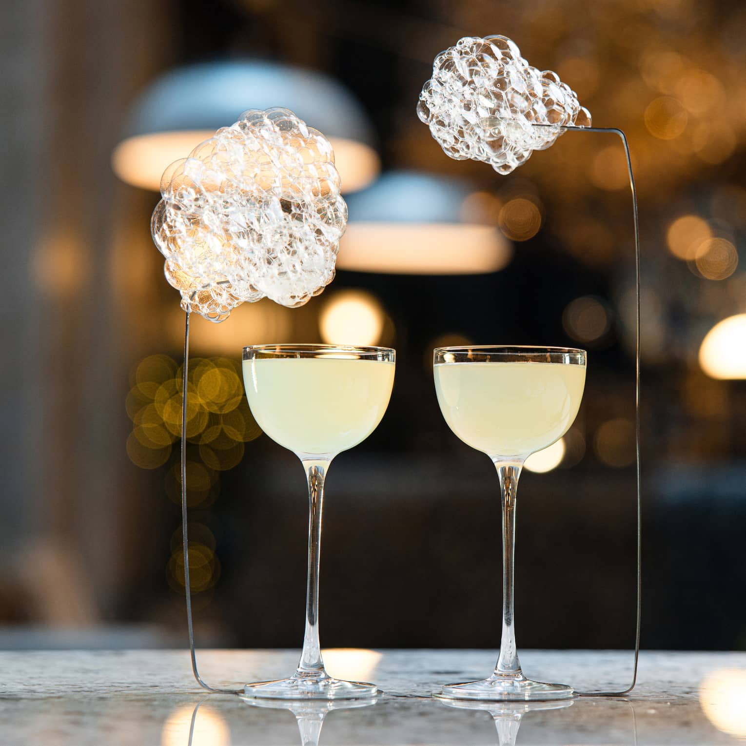 Two cloudy cocktails in unique glasses beside lamp on bar
