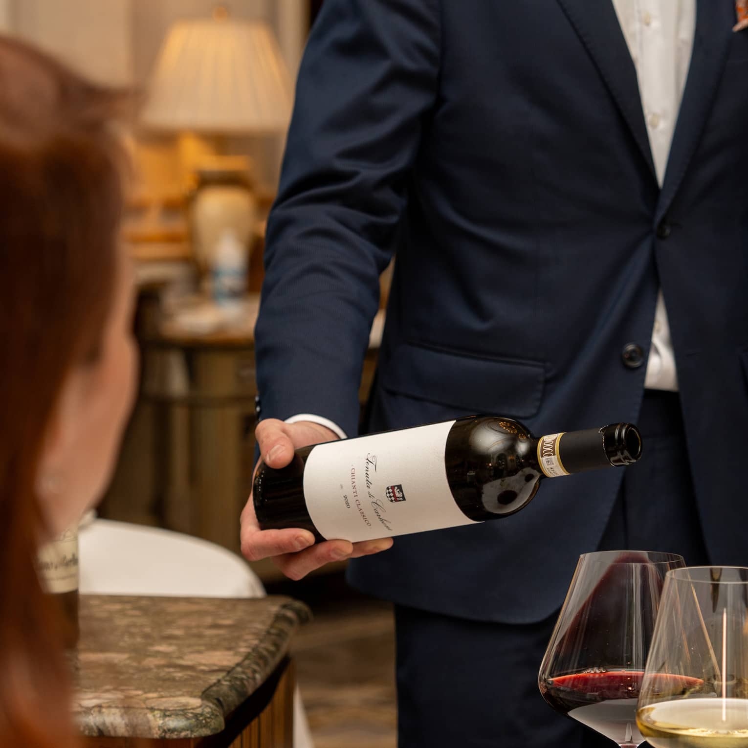 A guest sits at a table with three glasses of wine as a smiling sommelier tilts a bottle of red wine over one of the glasses.
