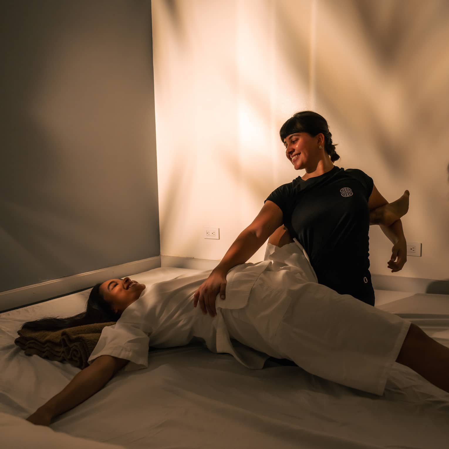 Two people sit in shadowy room as one of them helps stretch the back of the other