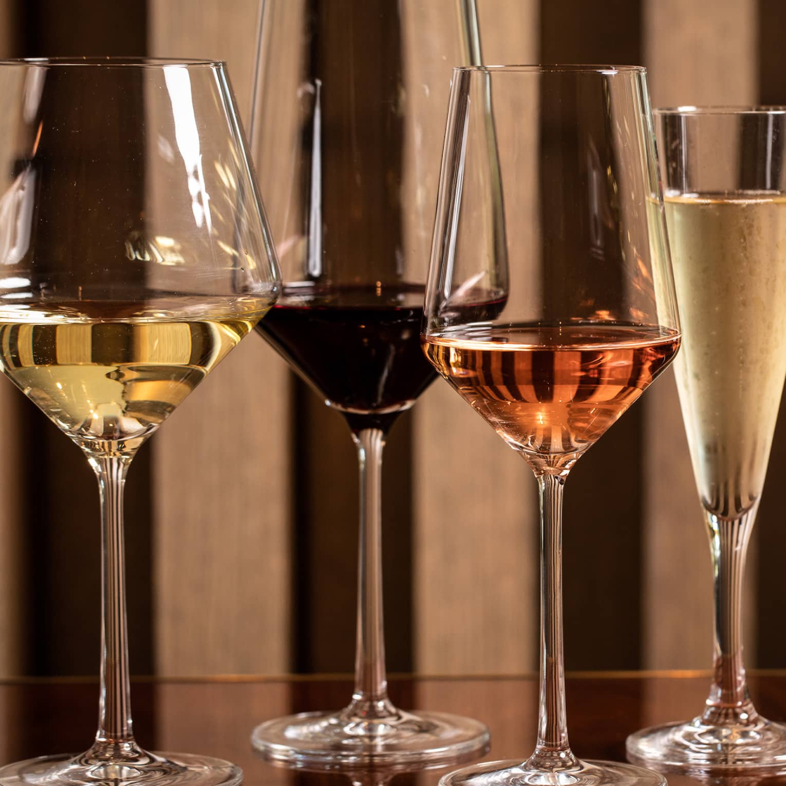 Four modern wine glasses holding white, red and rose wine are lined up on a wooden table