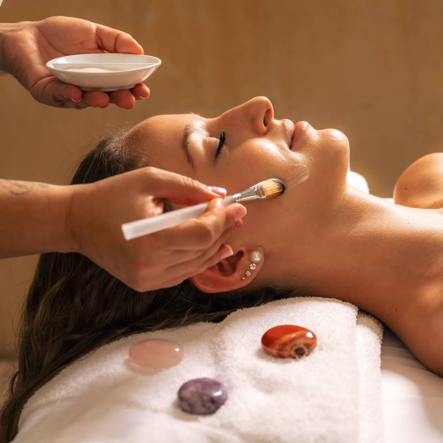 Woman lies covered by white blanket from shoulders down as someone gently applies a cream on their face with a brush, with three gemstones places next to the client's head