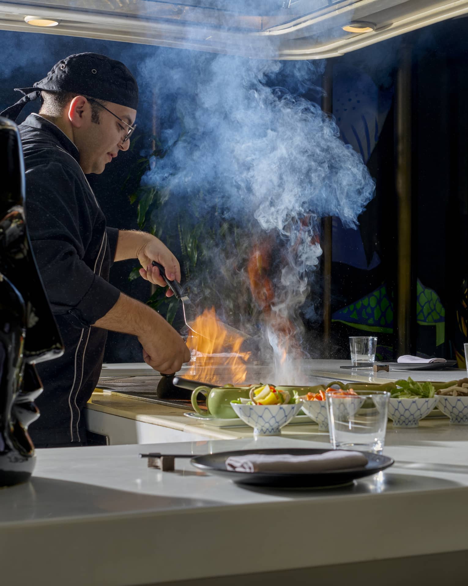Chef in black coat cooks dish over open flame in show kitchen