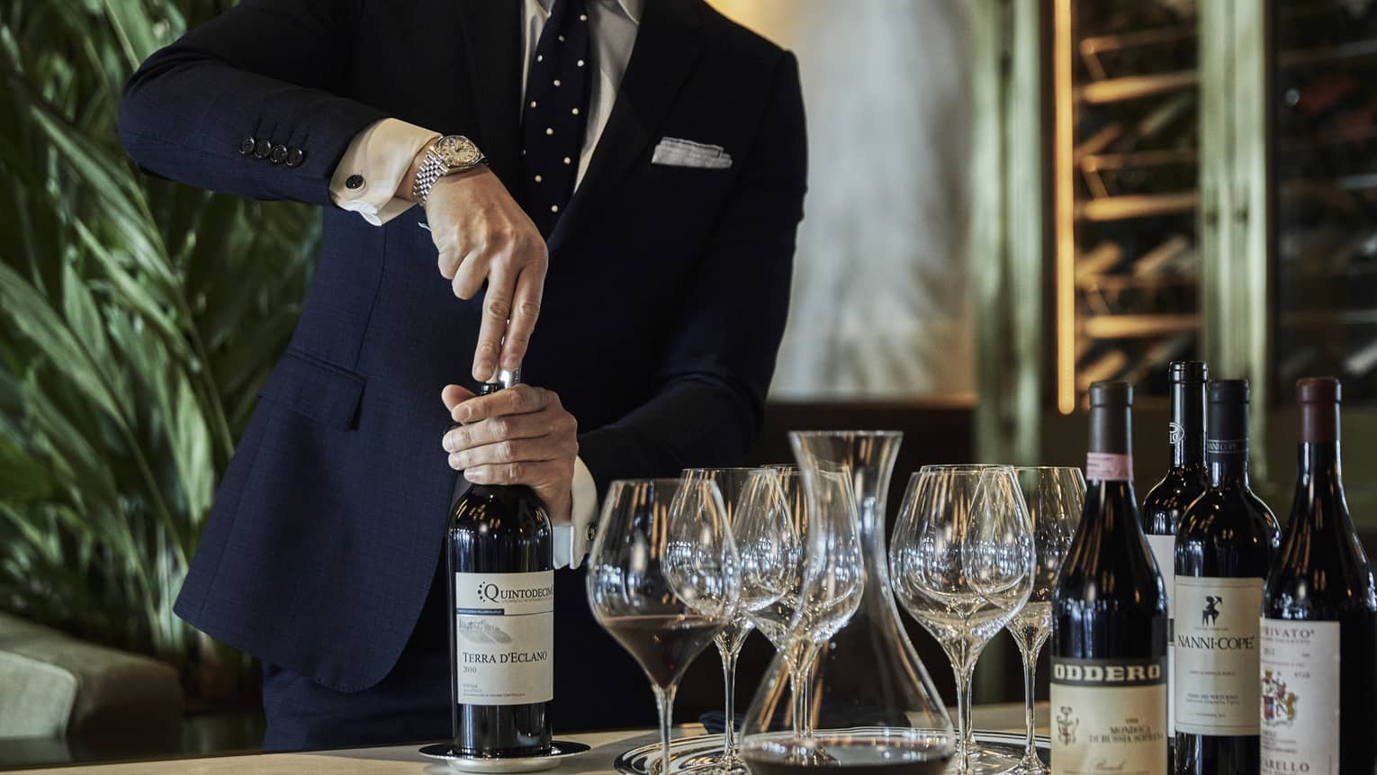 A person opening a bottle of wine, with wine glasses and additional wine bottles beside it