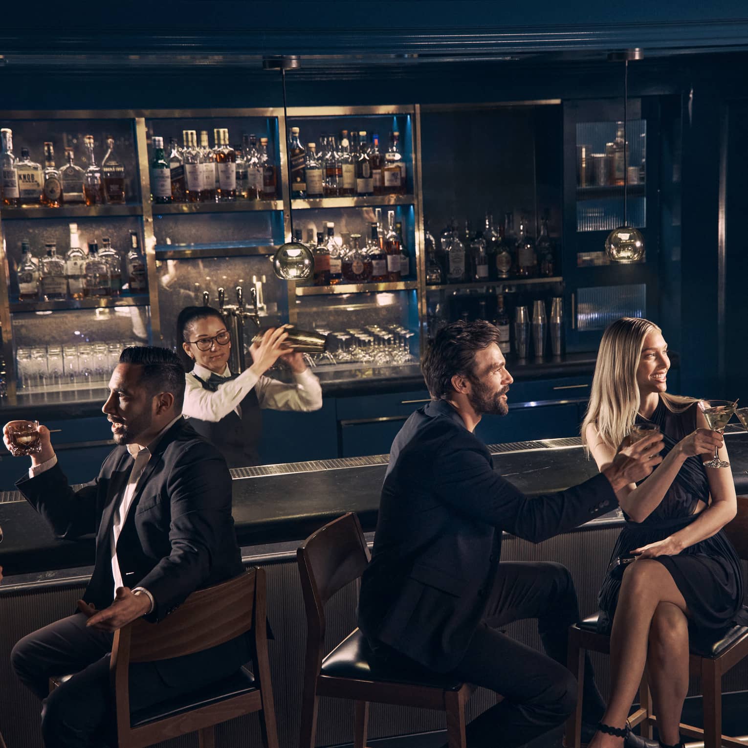 Guests chat over drinks at the bar at Prosperous Penny as a bartender shakes a cocktail shaker behind the bar.