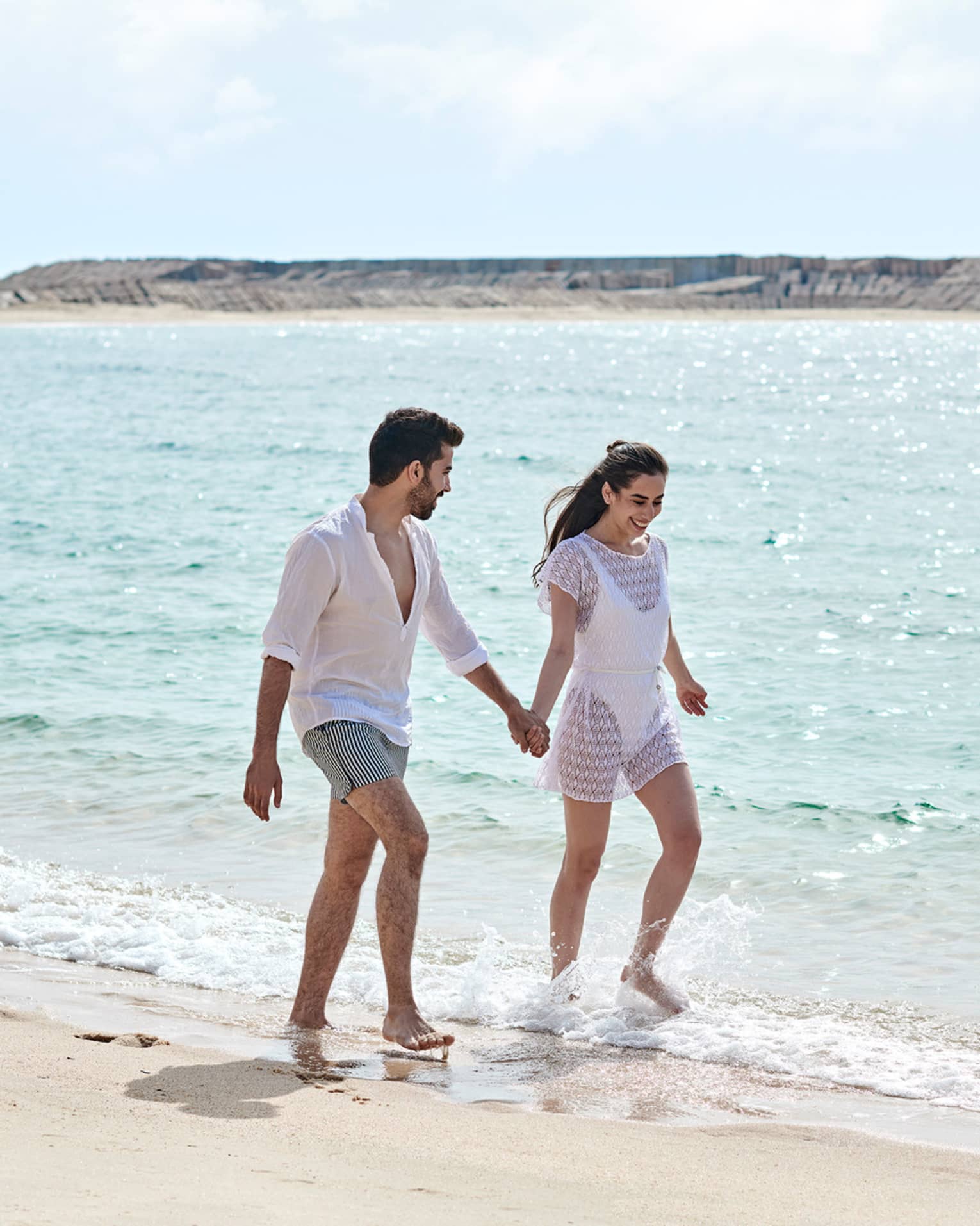 A man and woman both dressed in white hold hands and walk on a beach