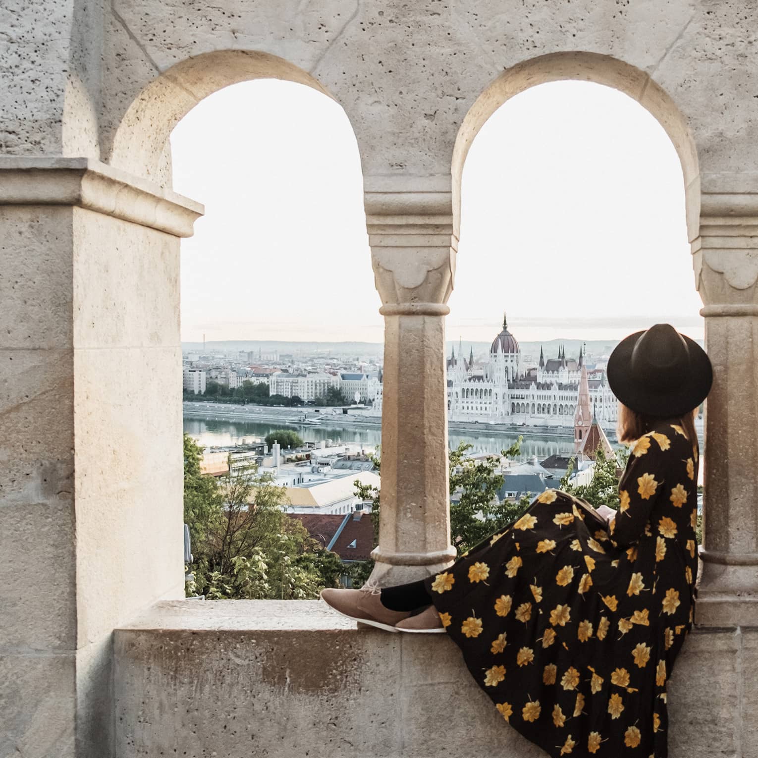 A guest in a floral dress and hat sitting between stone arches, gazing at a distant cityscape and river.