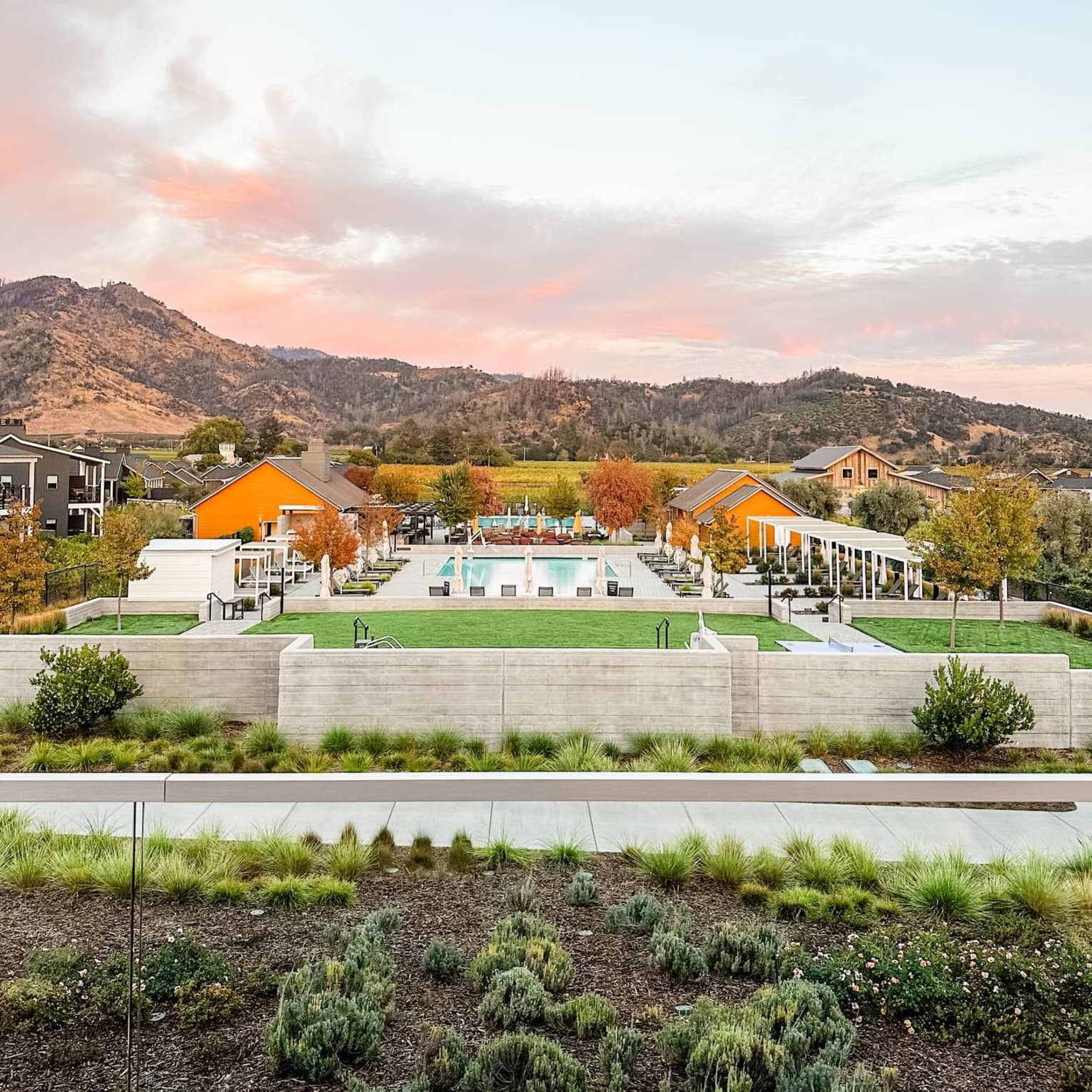 Resort pool area with surrounding greenery and buildings, set against a backdrop of rolling hills and a pink-tinged sunset sky.