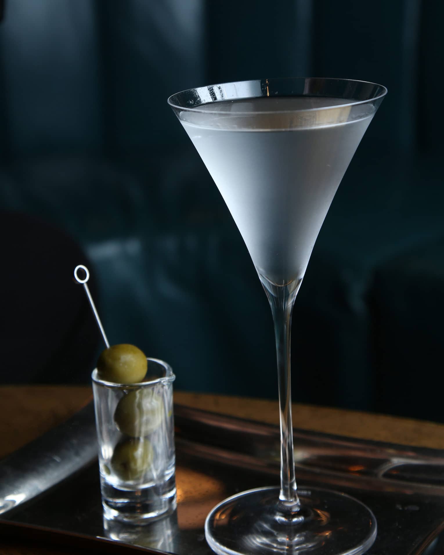 Chilled martini in a stemmed glass on a silver tray, accompanied by a small glass with green olives on a cocktail pick