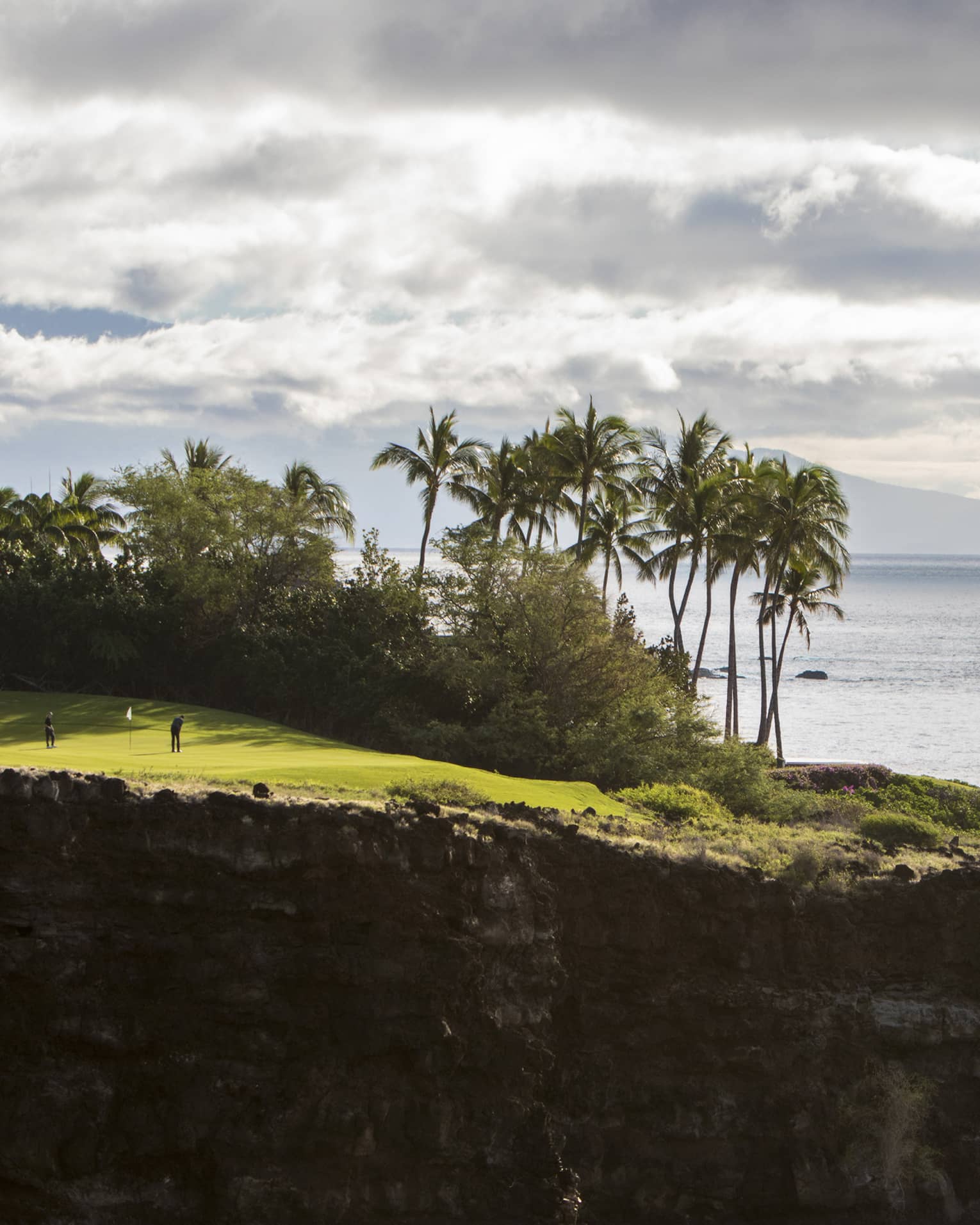 Long view of two people on clifftop golf green aside lush trees and towering palms; fluffy clouds, ocean and mountain beyond.