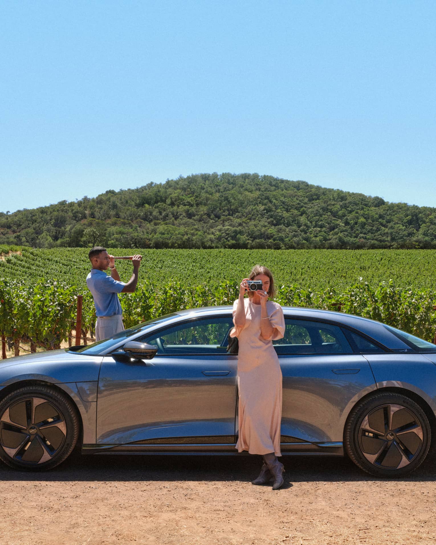 Person wearing long light-pink dress stands leaning against a grey car taking a picture of the surrounding vineyards with their camera while another person wearing a blue shirt and white pants stand on the other side of the car