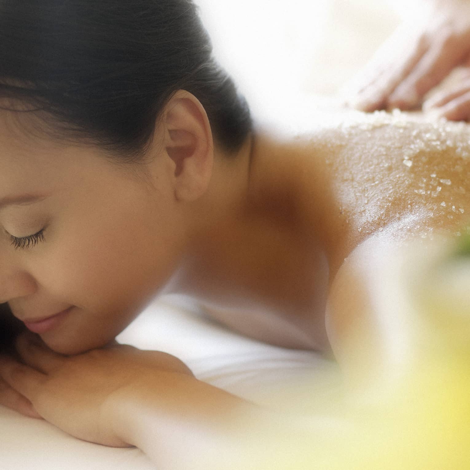Woman lying down with eyes closed as hands massage salt scrub into shoulders