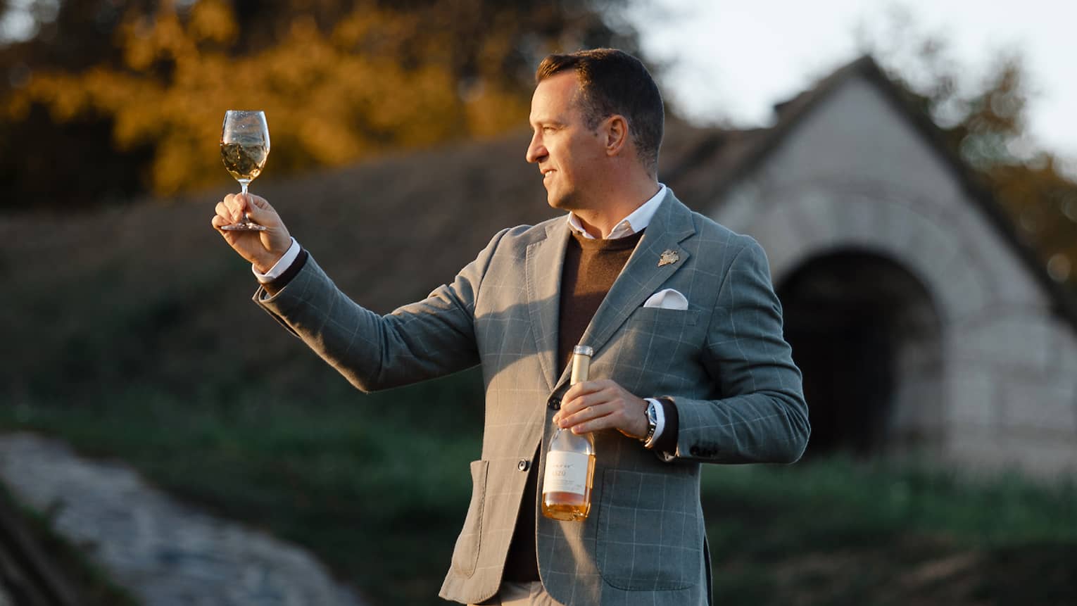 Person in a suit holding a wine bottle and inspecting a glass of white wine outdoors