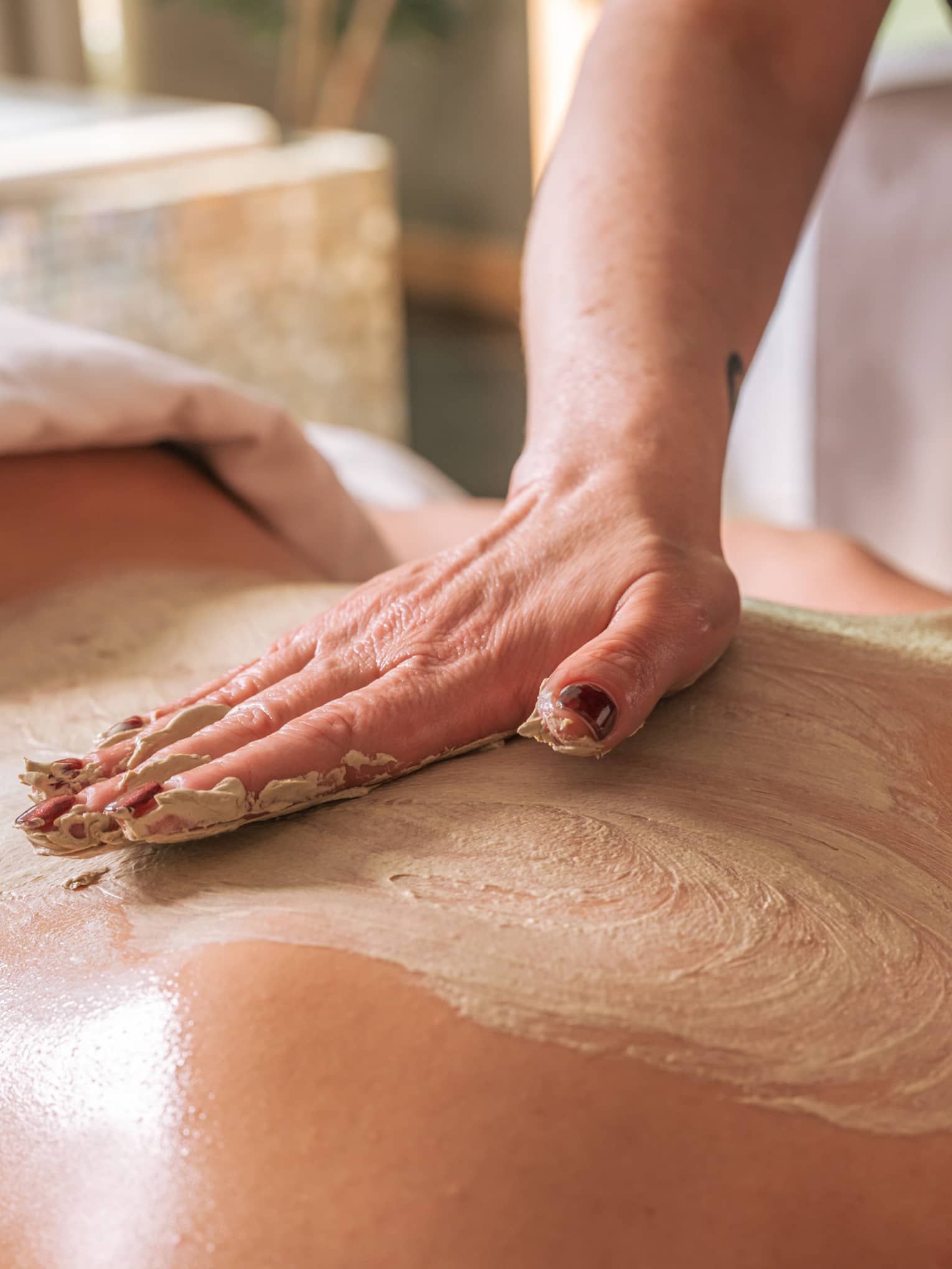 A hand rubs a sand-coloured body treatment on a person's bare back