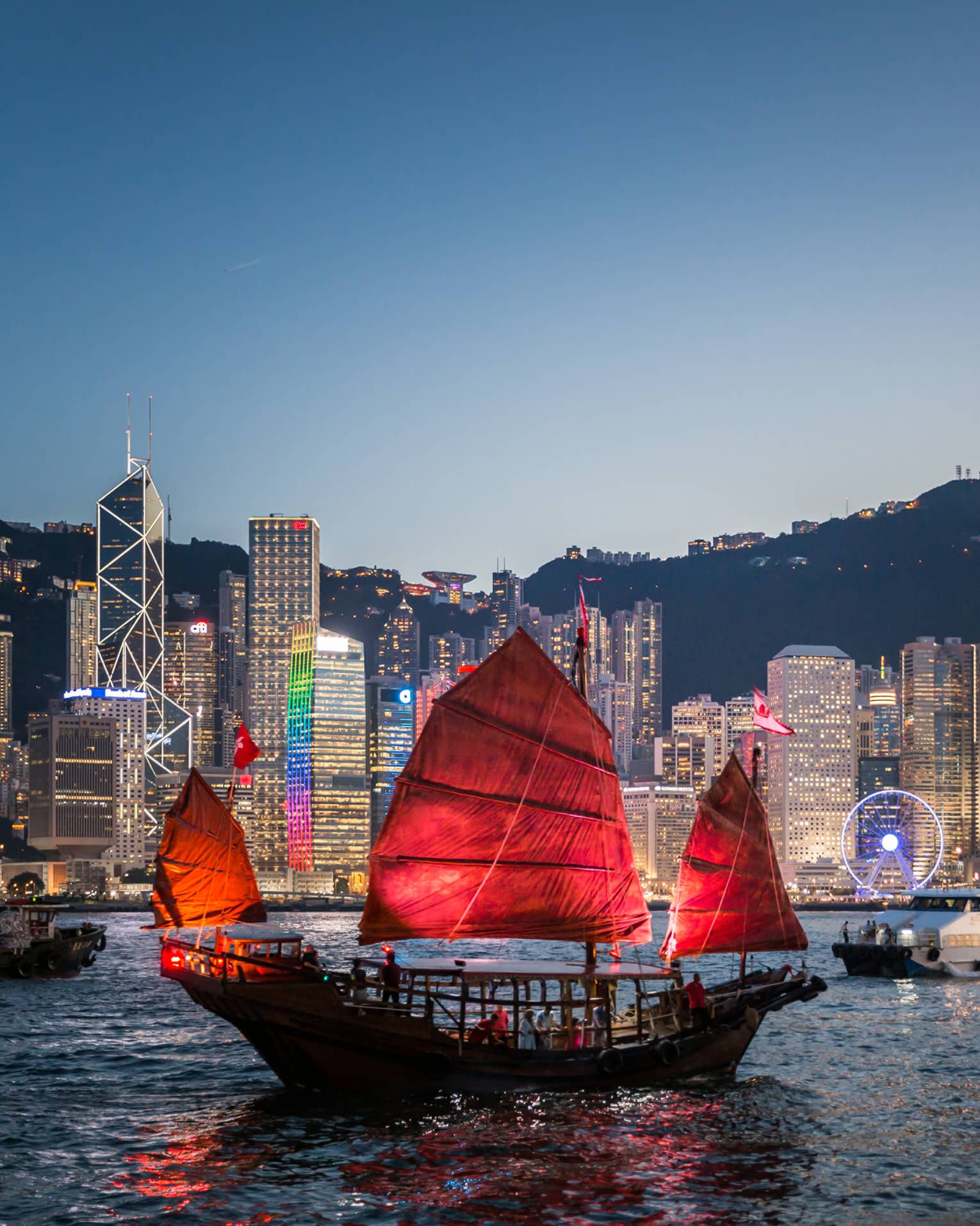 A traditional Hong Kong junk boat with red sails gliding through Victoria Harbour at dusk, with the city’s illuminated skyline in the background