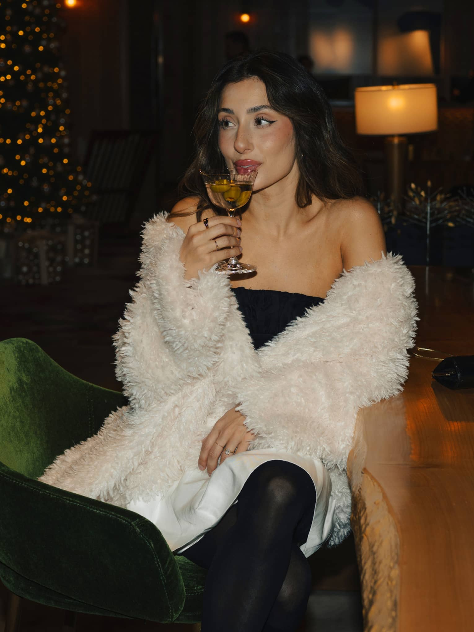 Person with long dark hair wears a dark-green strapless dress and white faux fur coat while sipping a cocktail