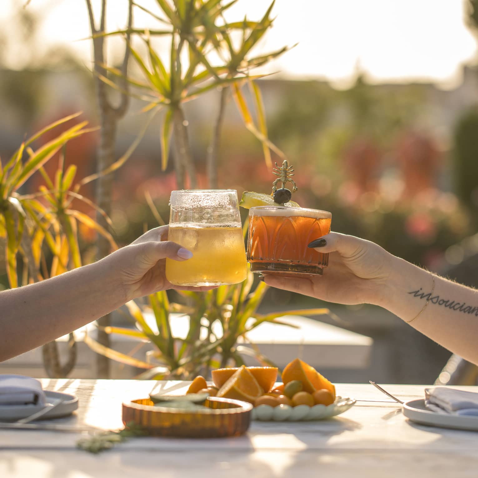 Two hands, each holding a cocktail, bring their glasses together over a sun-lit outdoor table