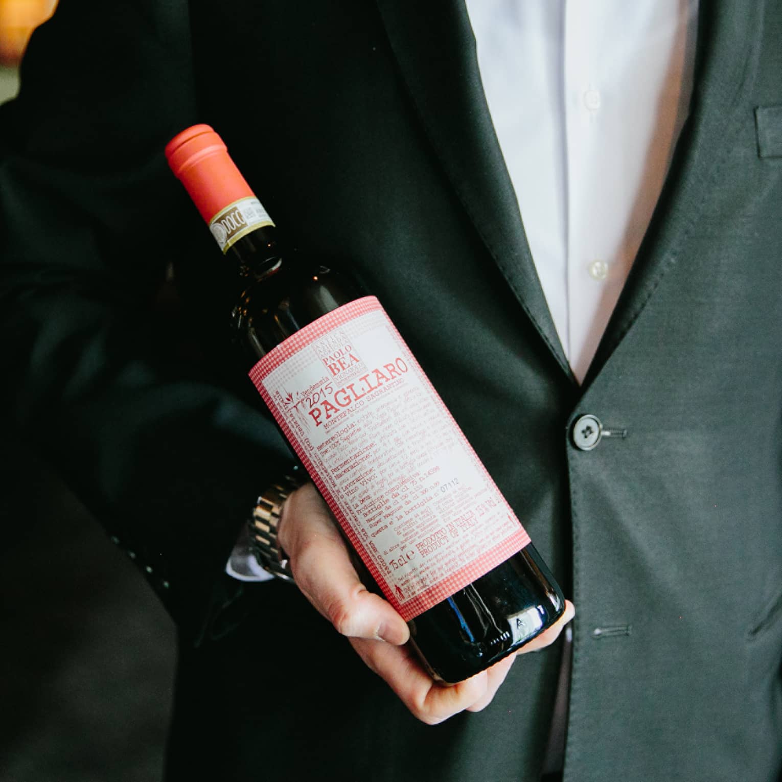 A man dressed in a suit holds a bottle of wine with a red label