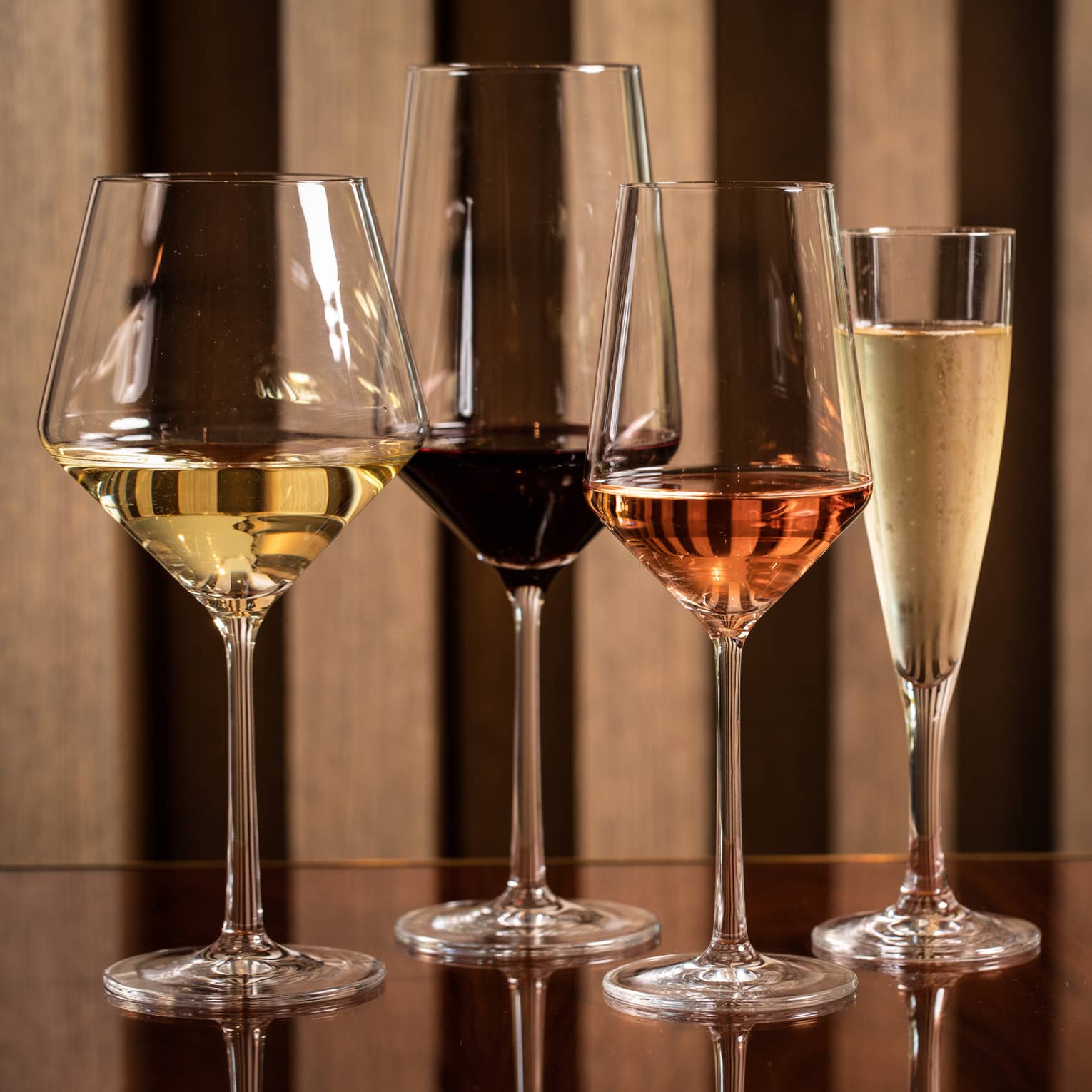 Four modern wine glasses holding white, red and rose wine are lined up on a wooden table