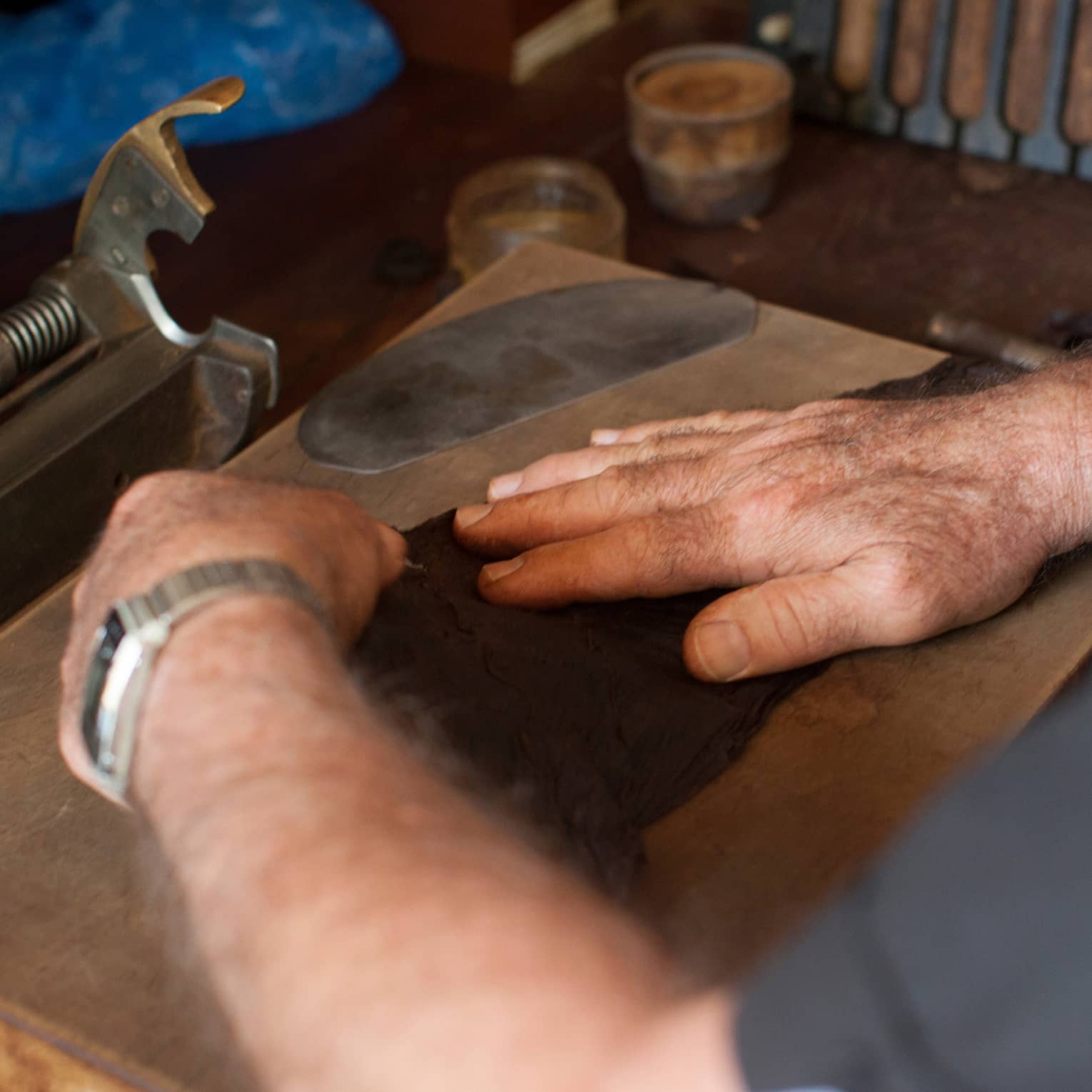 Close-up of hands rolling a flat cigar leaf on a wooden board, near a metal cigar cutter and a rack of completed cigars.