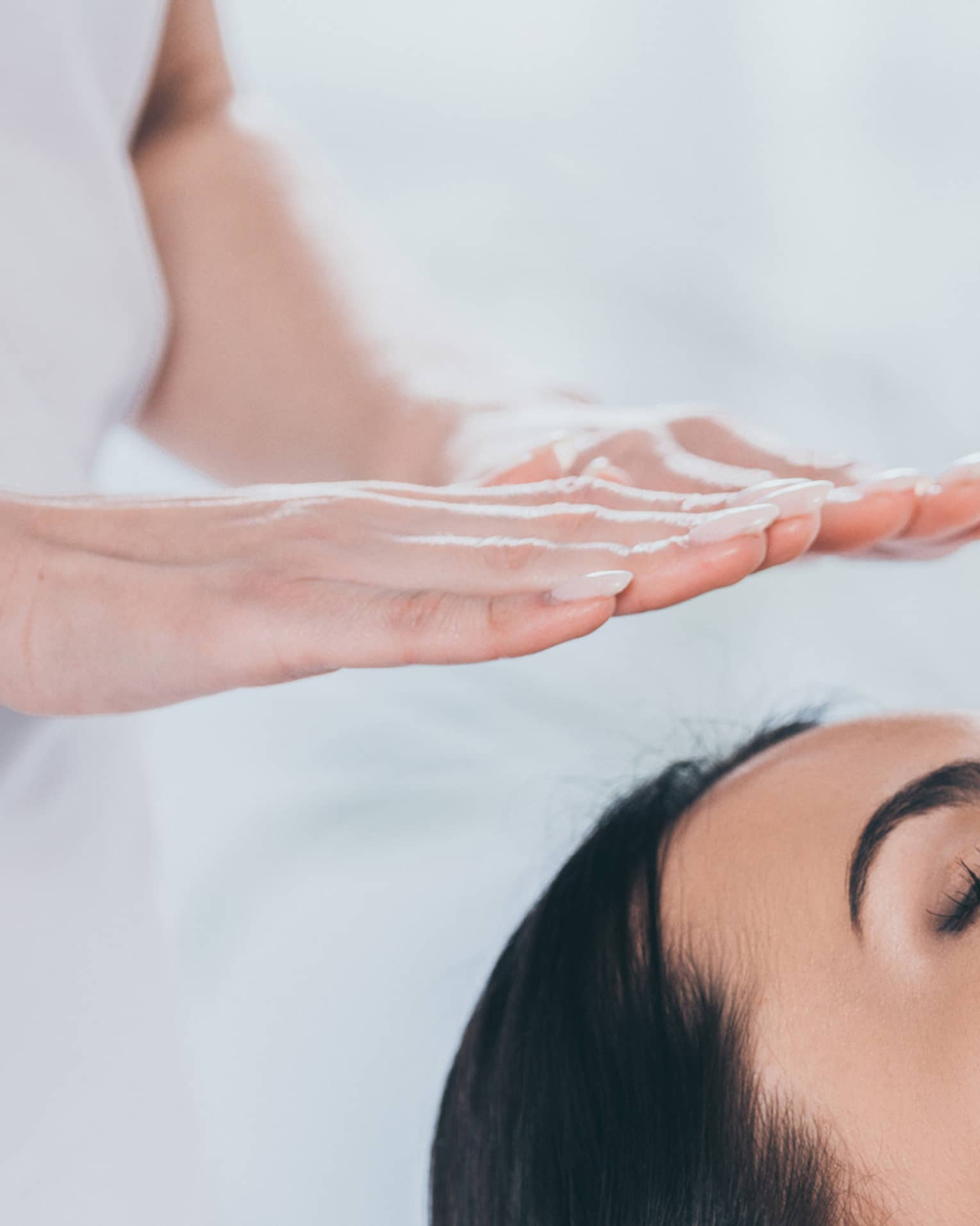 Woman with eyes closed lays on rolled up towel on spa table under therapists' hovering hands