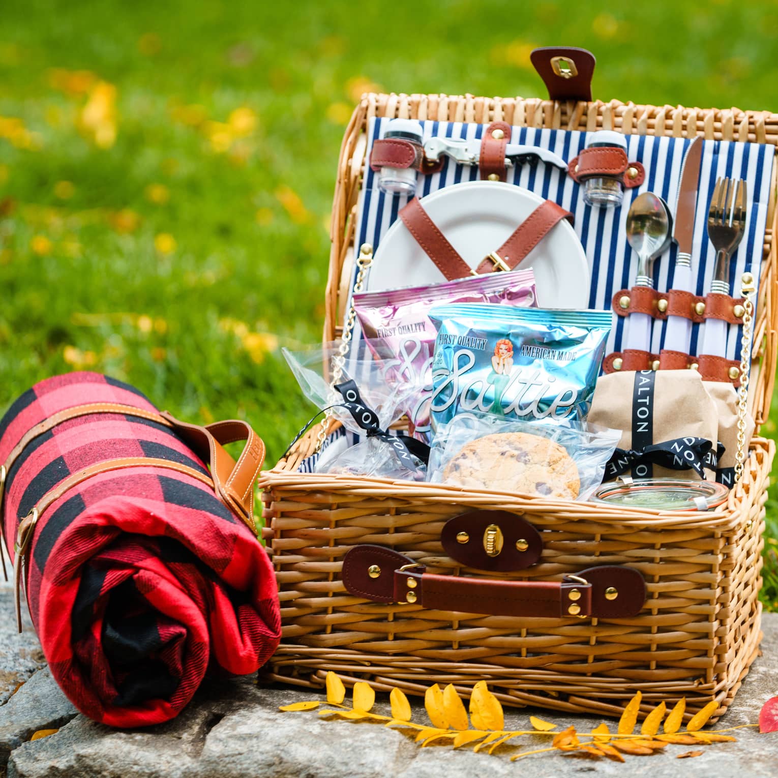 A picnic basket with plates, utensils and snacks resting on a stone near grass and yellow leaves.
